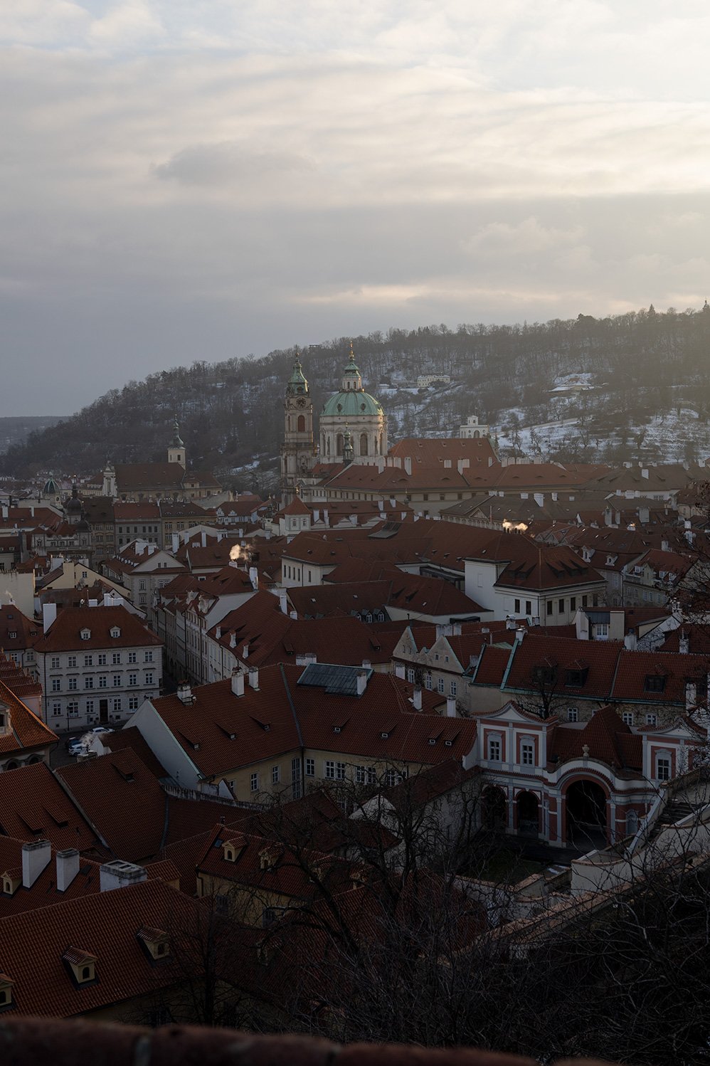 A cityscape with red rooftops, a prominent cathedral with a green dome, and snow-covered hills in the background under a cloudy sky.