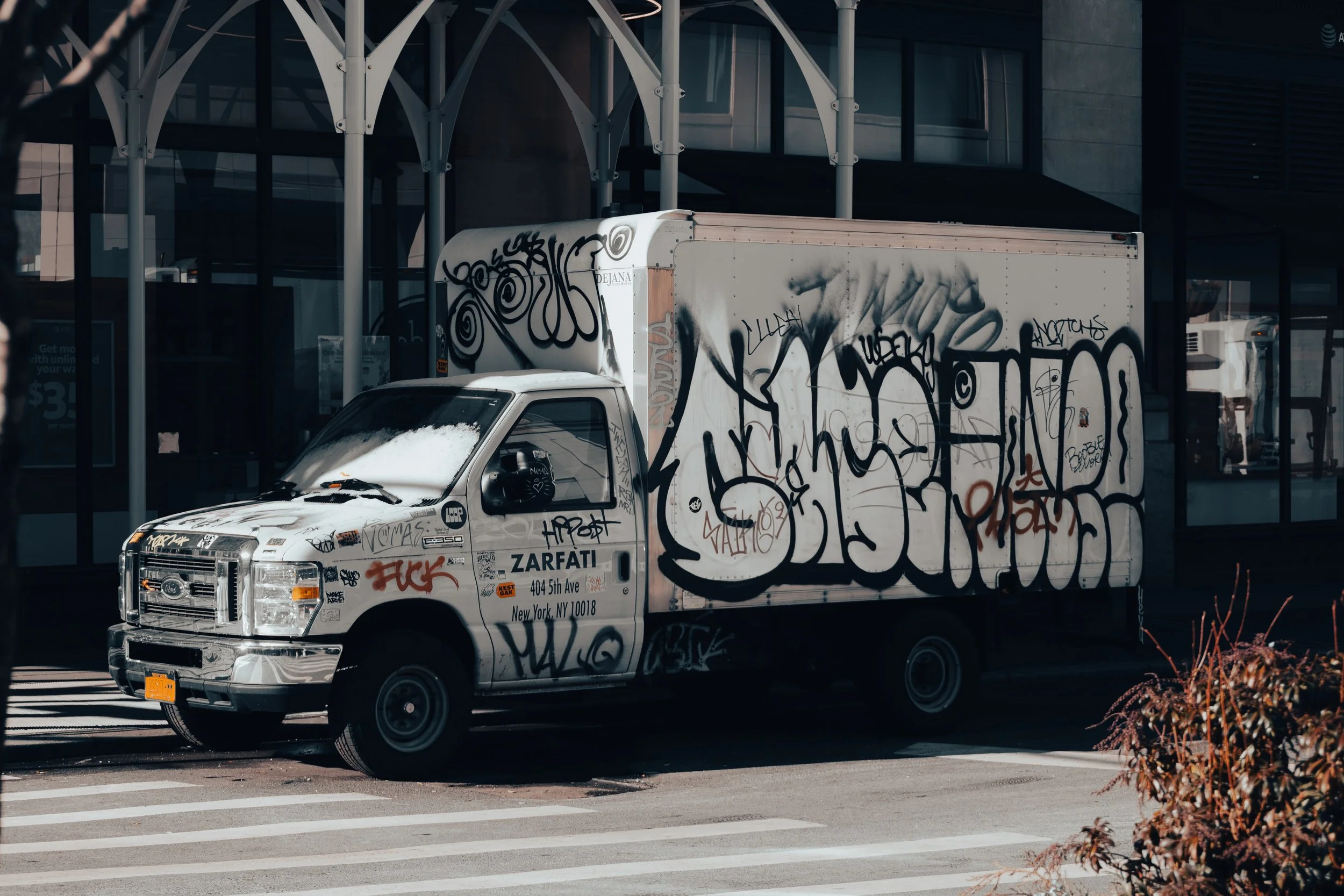 A white delivery truck covered in black graffiti parked on the side of the street in front of a modern glass building.