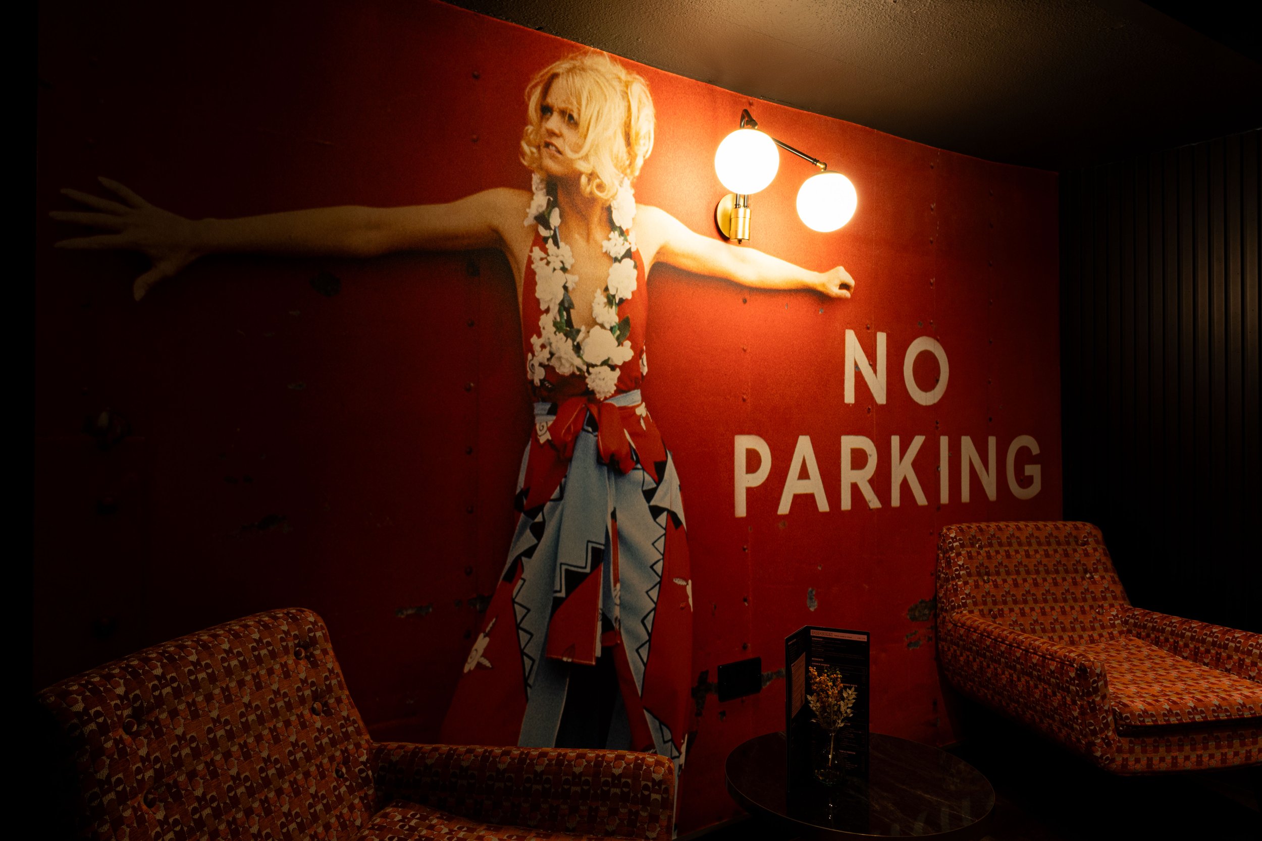 A poster of a woman with blonde hair in a floral dress with arms outstretched mounted on a red wall, with two round light fixtures above her, and the partial text reading 'NO PARKING' on the wall, in a room with patterned chairs.