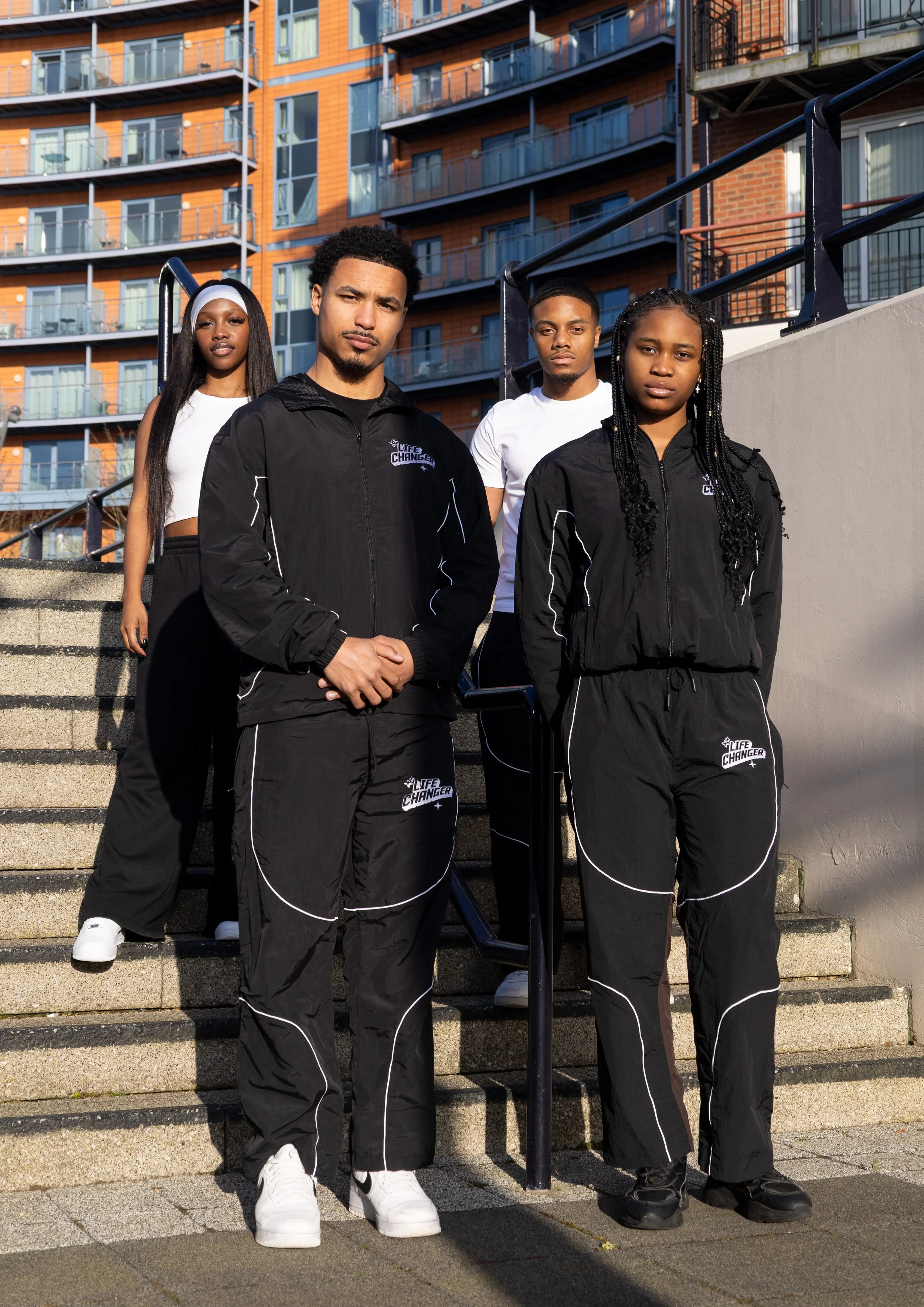 Group of four young adults standing outdoors on concrete steps in front of a modern apartment building, dressed in black and white athletic clothing with 'LIFE CHANGER' logos.