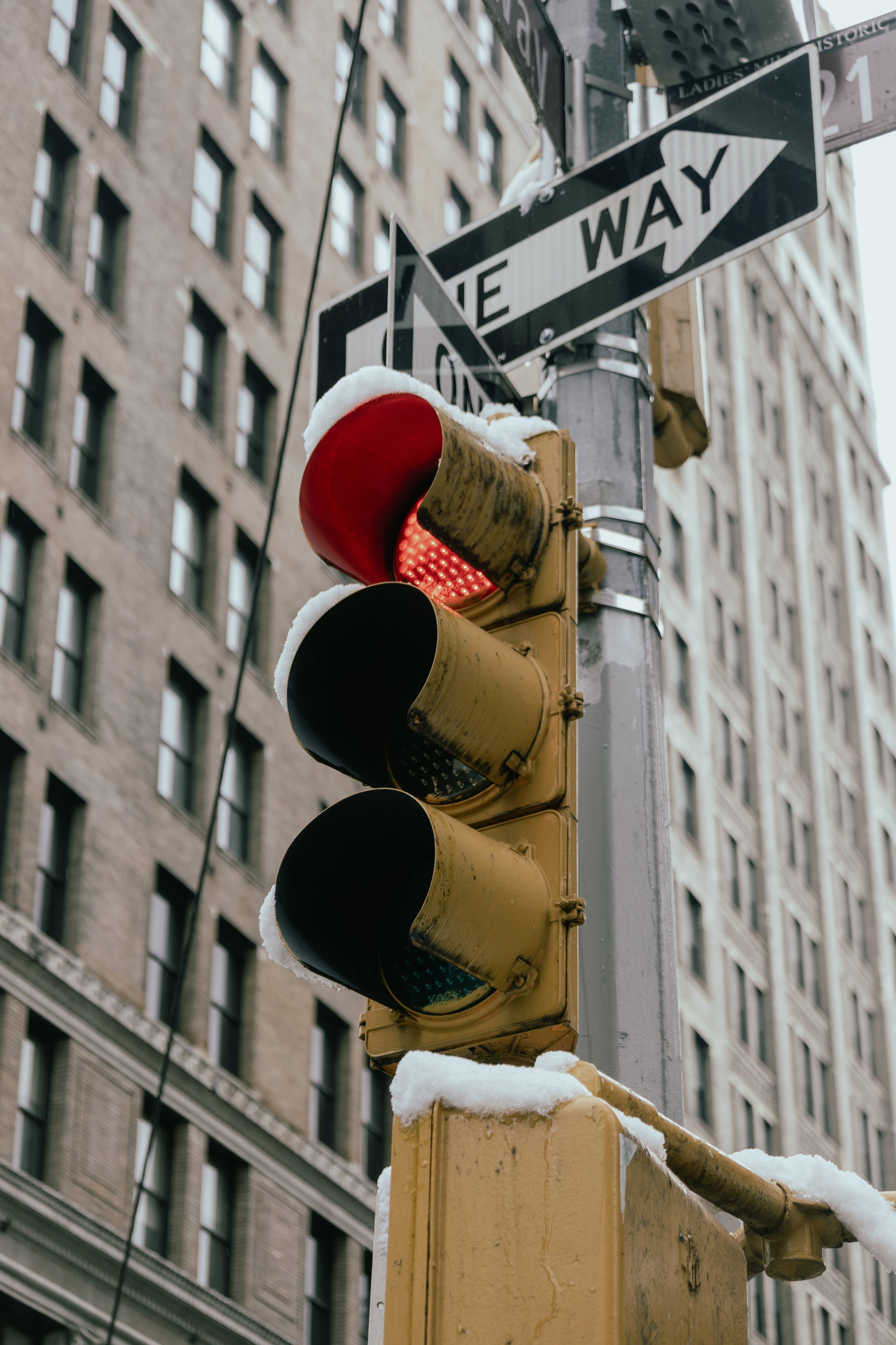 A traffic light showing red, with snow on it, and a street sign indicating one way, against a backdrop of tall buildings.