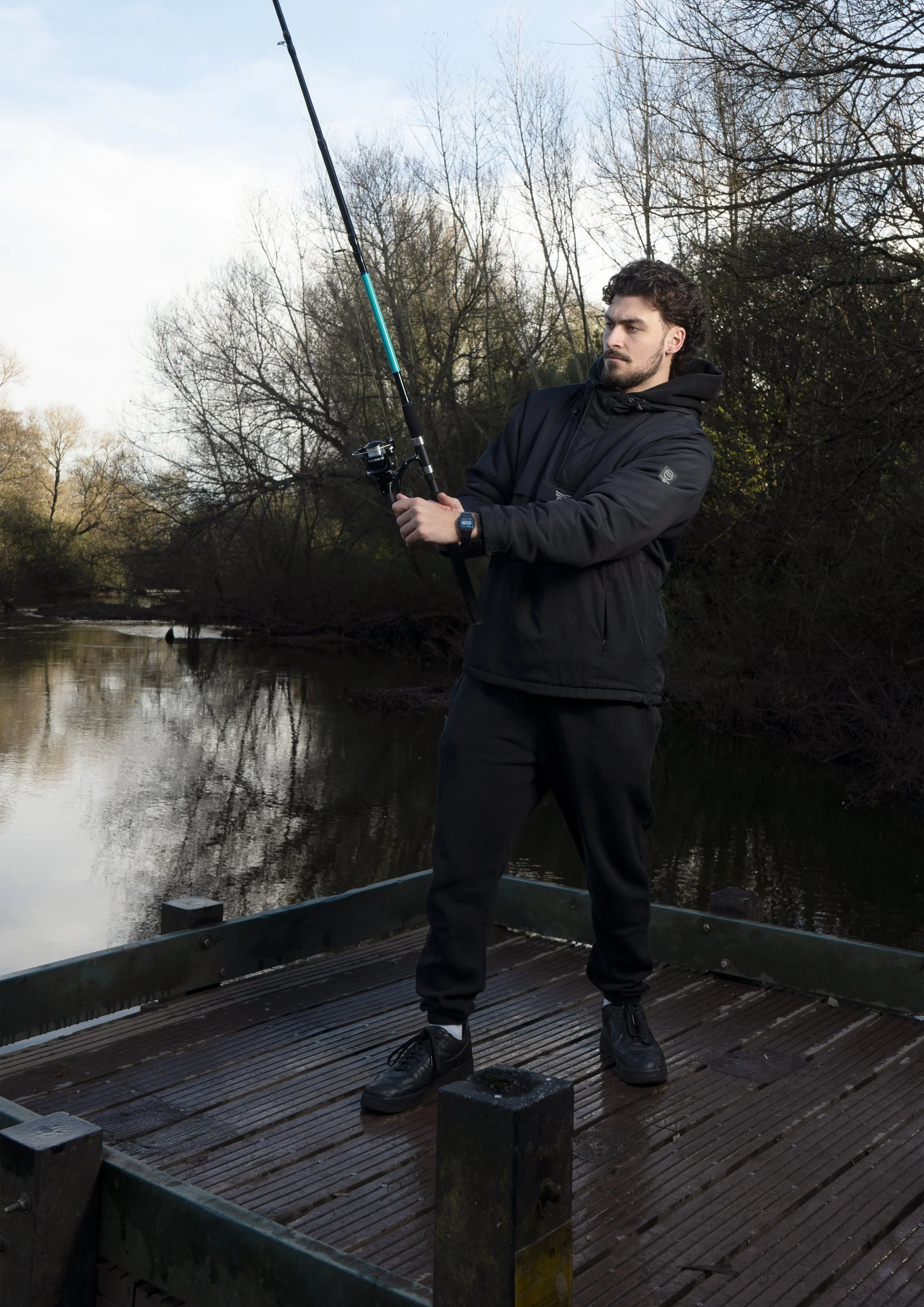 A man fishing on a river, standing on a wooden dock, holding a fishing rod, with trees and water in the background on a cloudy day.