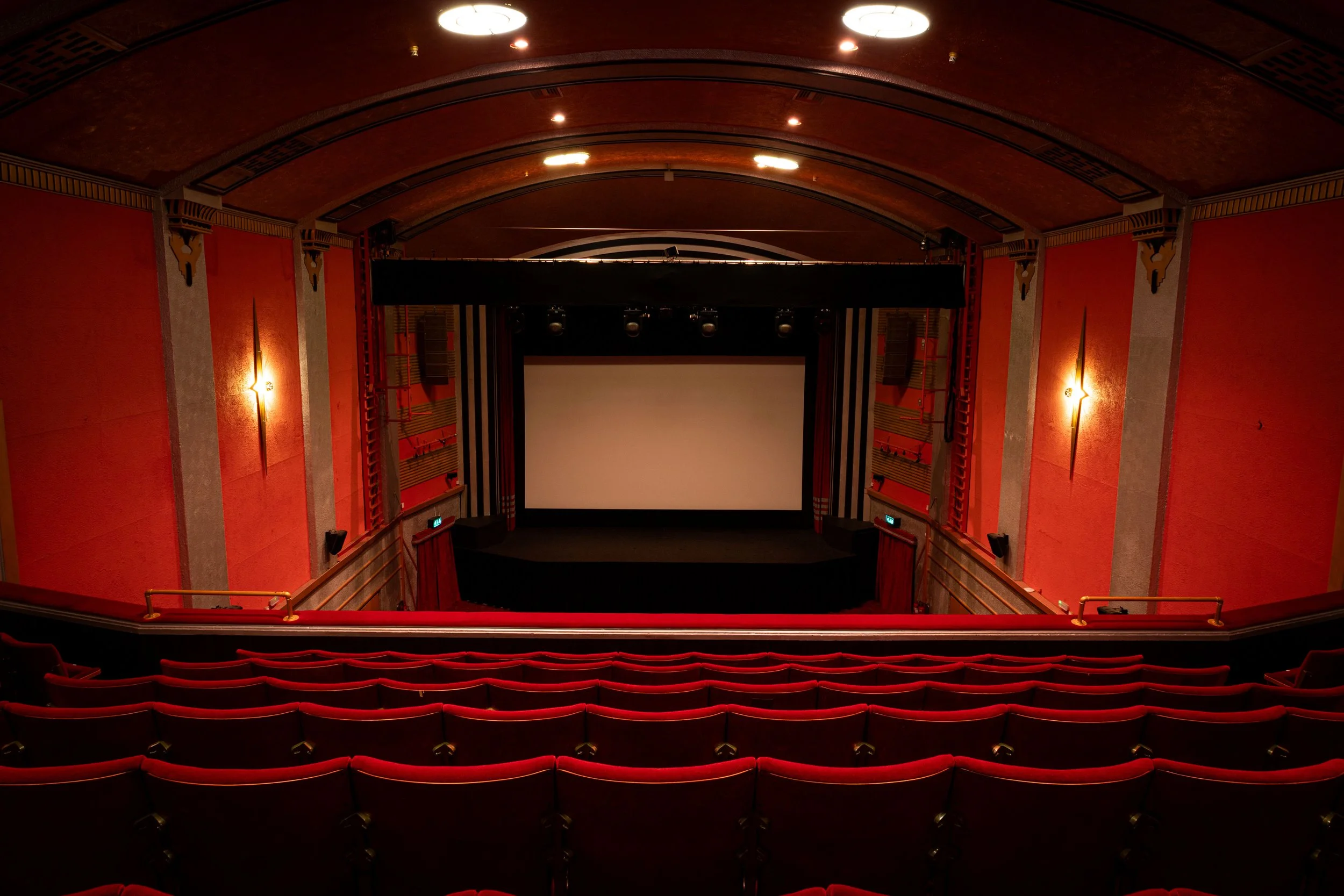 The interior of an empty movie theater with red seats, a large screen, and dim lighting.