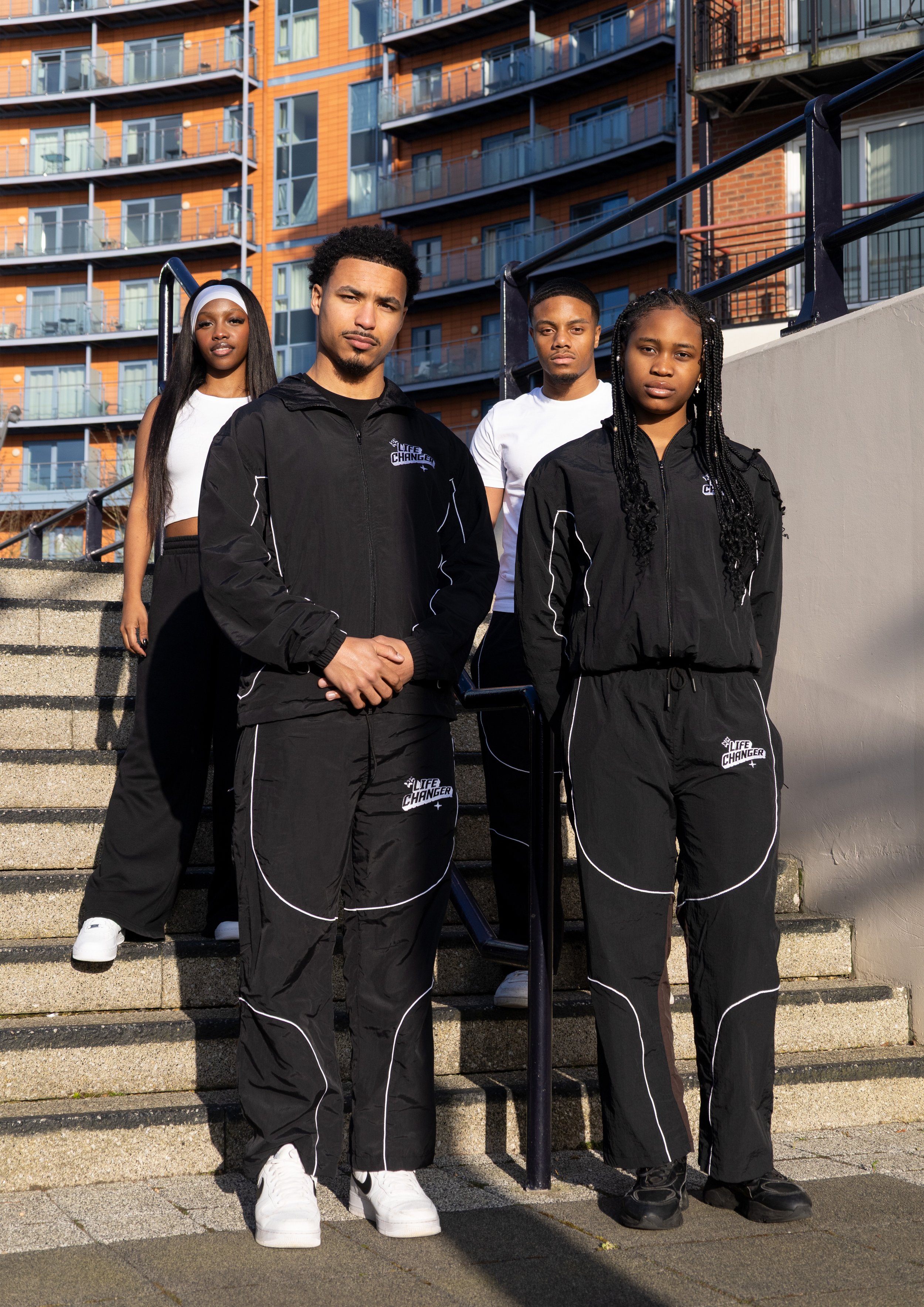 Four young adults standing on outdoor concrete stairs in front of an urban apartment building with orange exterior tiles and glass balconies. They wear black athletic tracksuits with white piping that have a logo reading 'LIFE CHANGER' and look serio