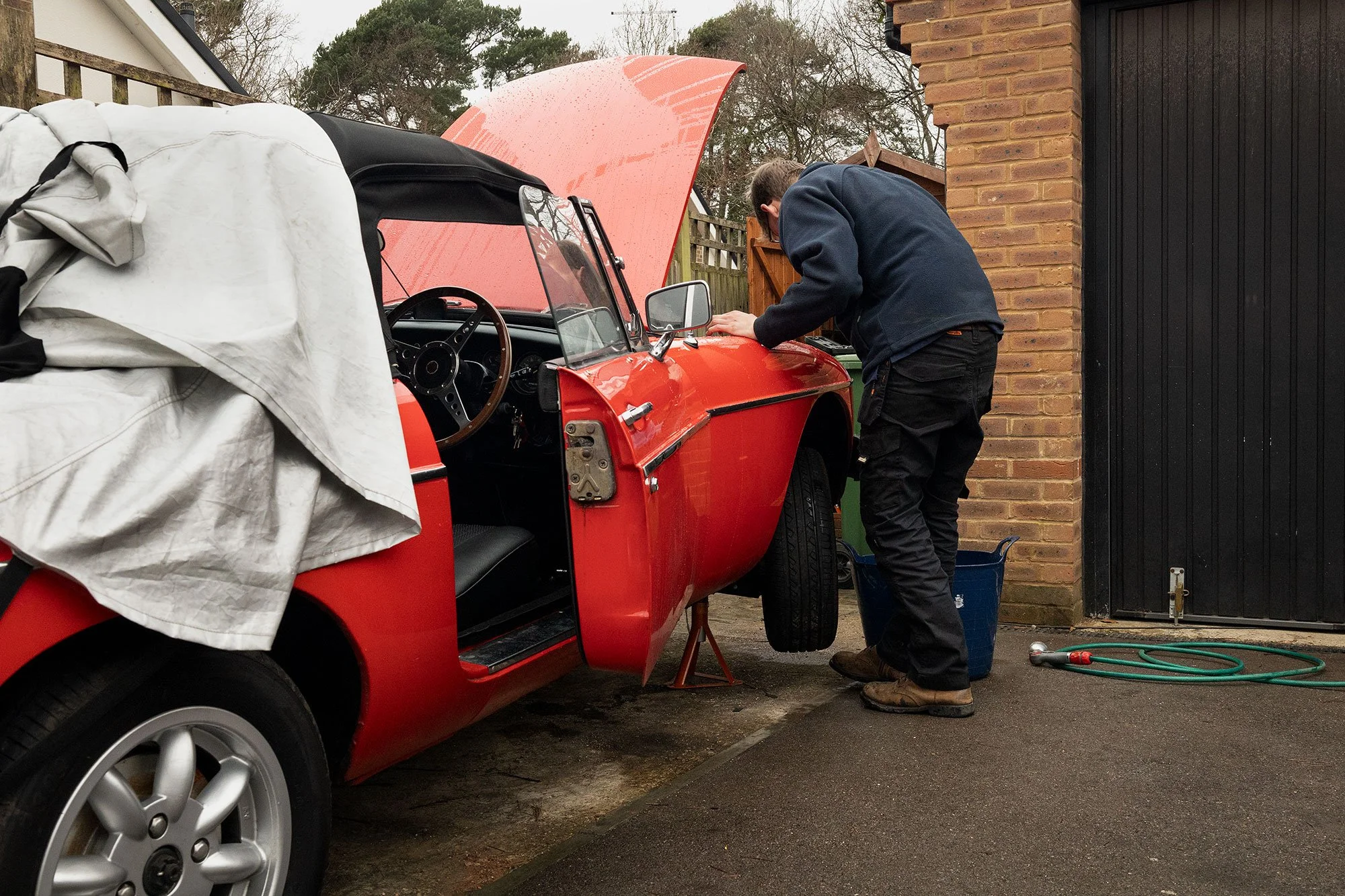 A man working on the engine of a red convertible car parked outside a house. The convertible has a partially open hood, and the man is leaning over the engine bay, wearing a navy jacket and black pants. There are gardening tools and a green garden ho
