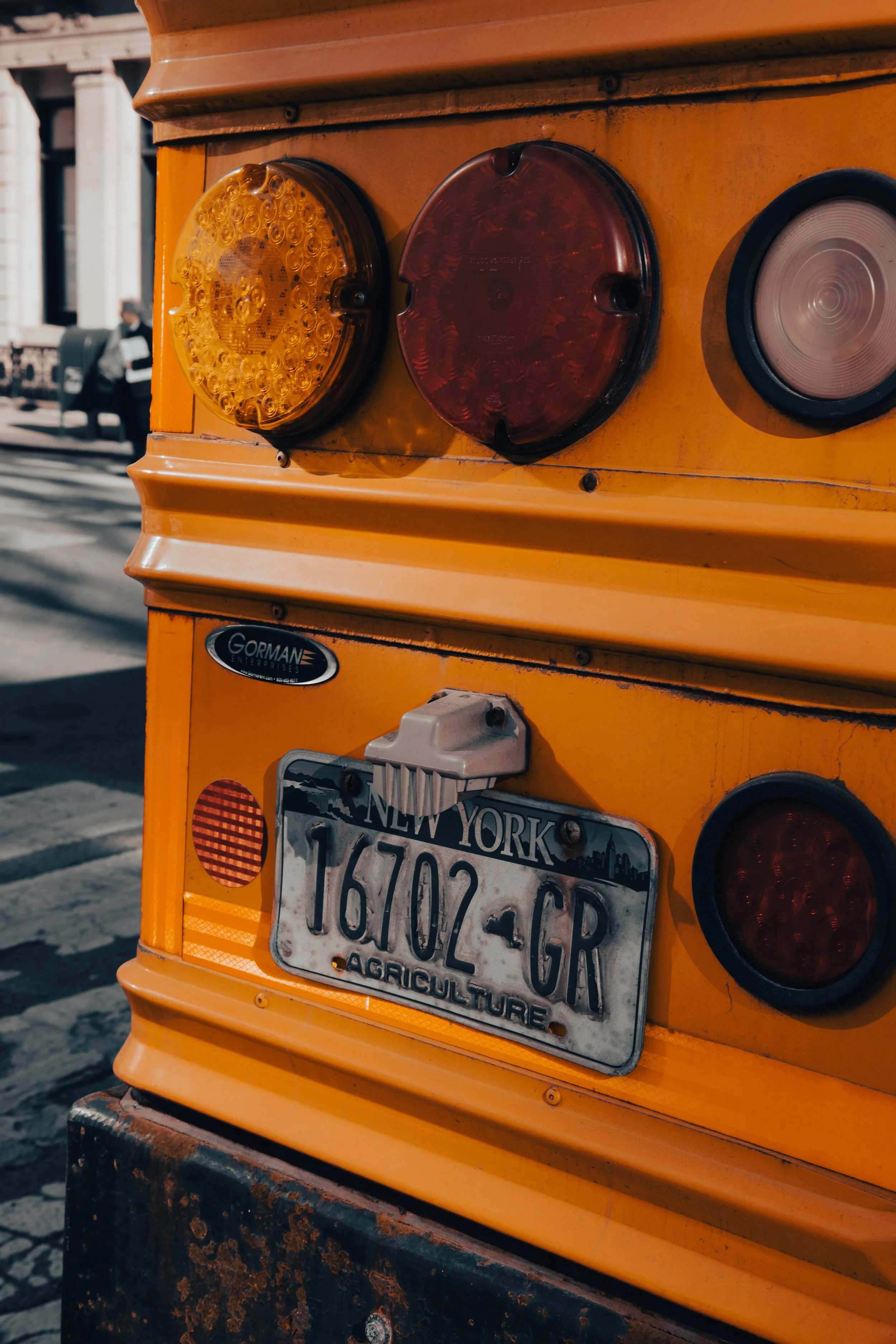 Close-up of a yellow utility truck with a New York license plate, showing round amber and red tail lights and a rusty black bumper in an urban setting.