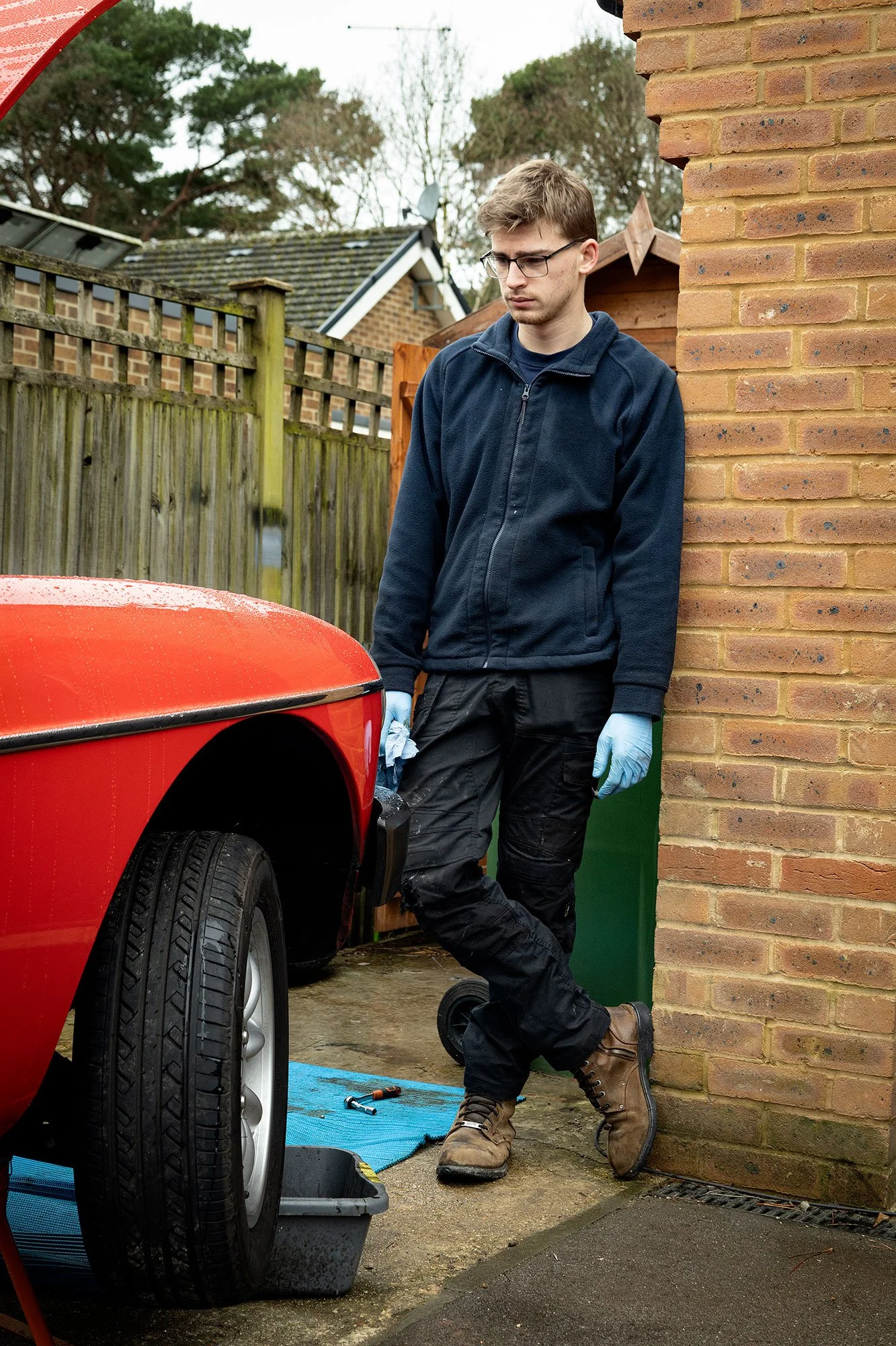 A young man with glasses and a navy jacket working on a car in a driveway, standing next to a brick wall, with tools and a blue mat on the ground.