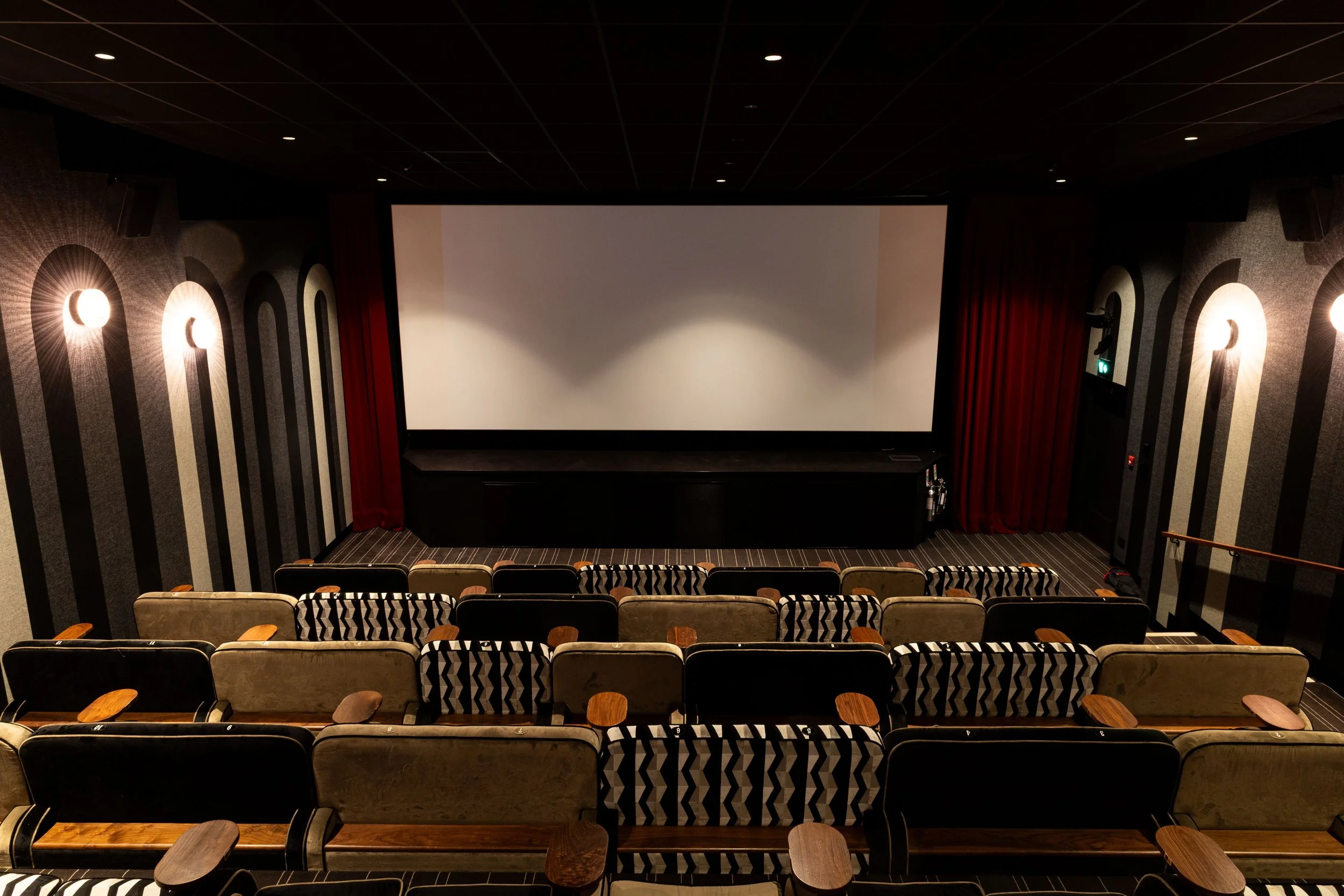 An empty movie theater with rows of patterned seats facing a large white screen and red curtains on either side.