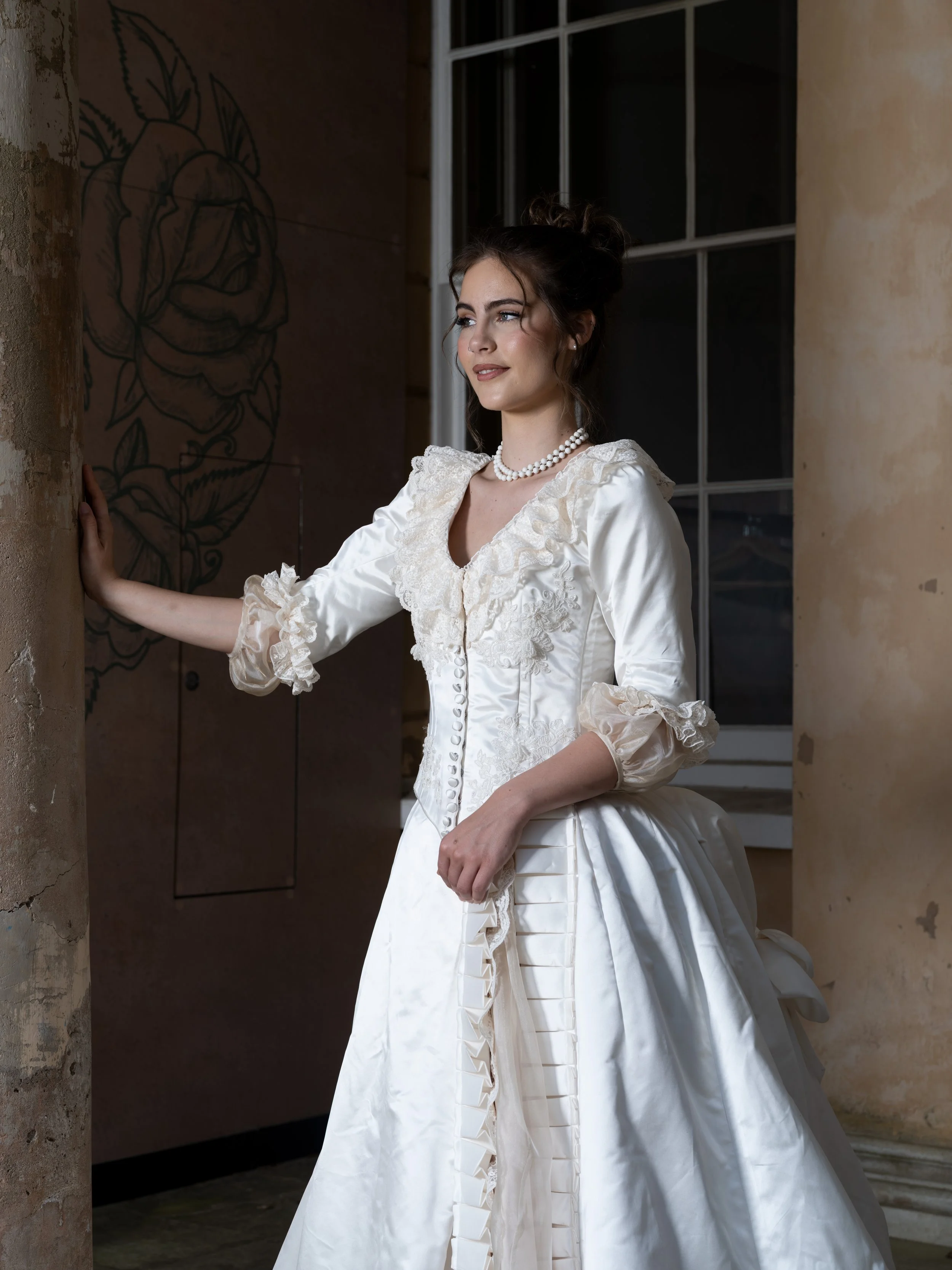 A woman dressed in an elegant vintage-style wedding dress with lace details and puffy sleeves, standing indoors near a wall with a painted floral design. She has dark hair styled in an updo and is wearing a pearl necklace, with a calm expression on h