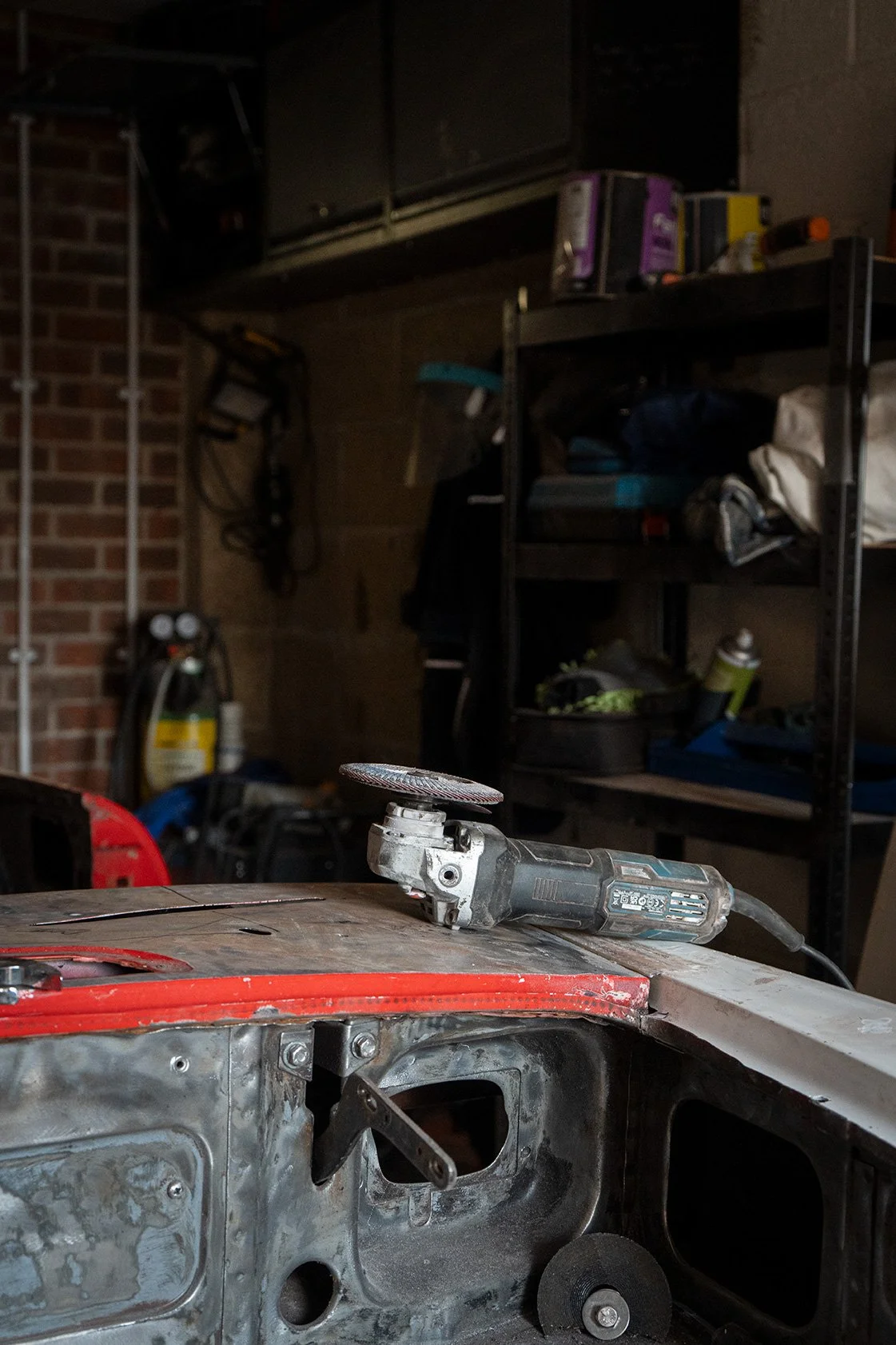 A garage workshop with a power grinder on a metal car body, tools, shelves with various supplies, and a brick wall in the background.