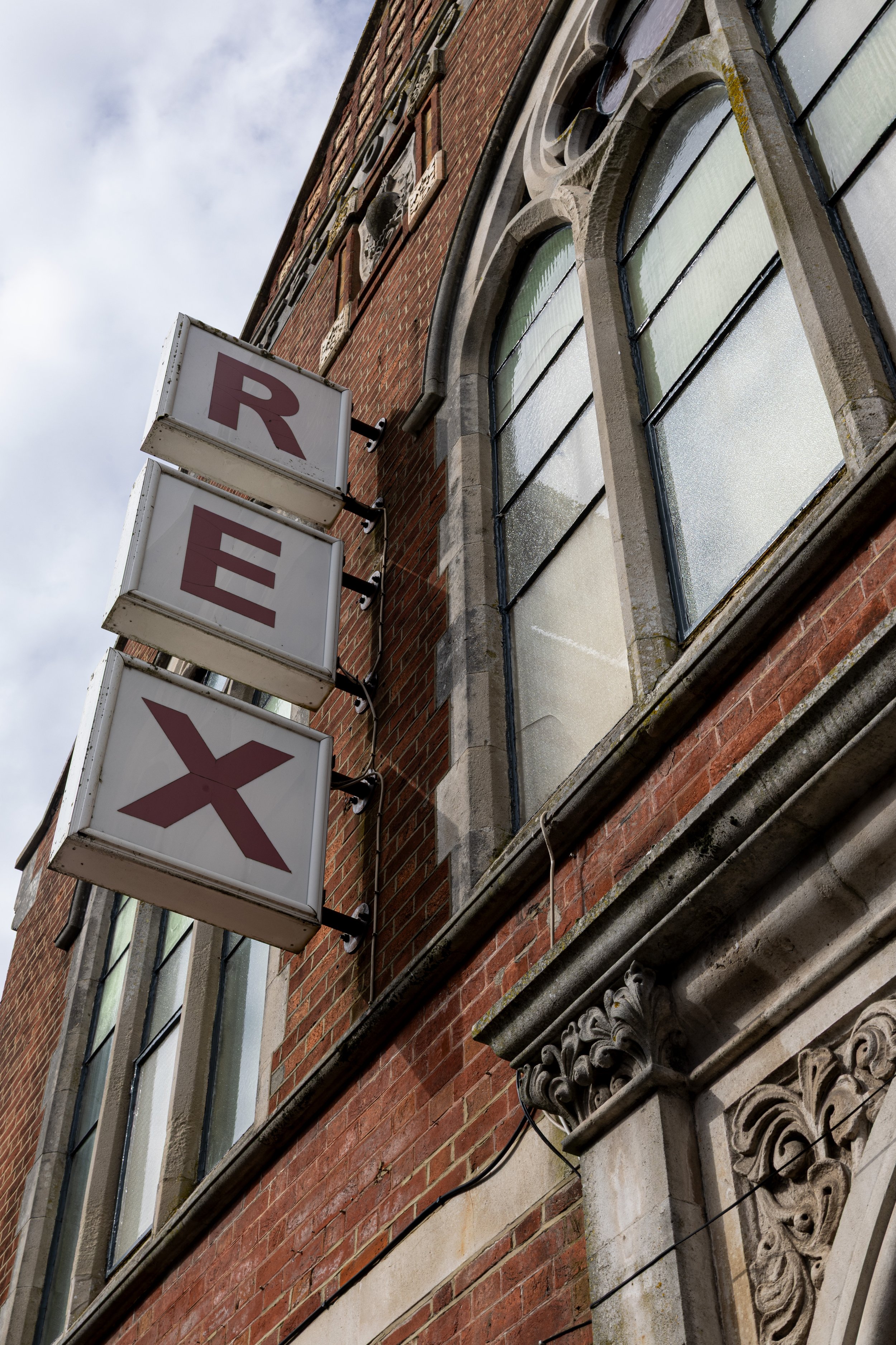 Close-up of a brick building facade with large arched windows and decorative stonework, featuring a vertical Sign with red letters on a white background.