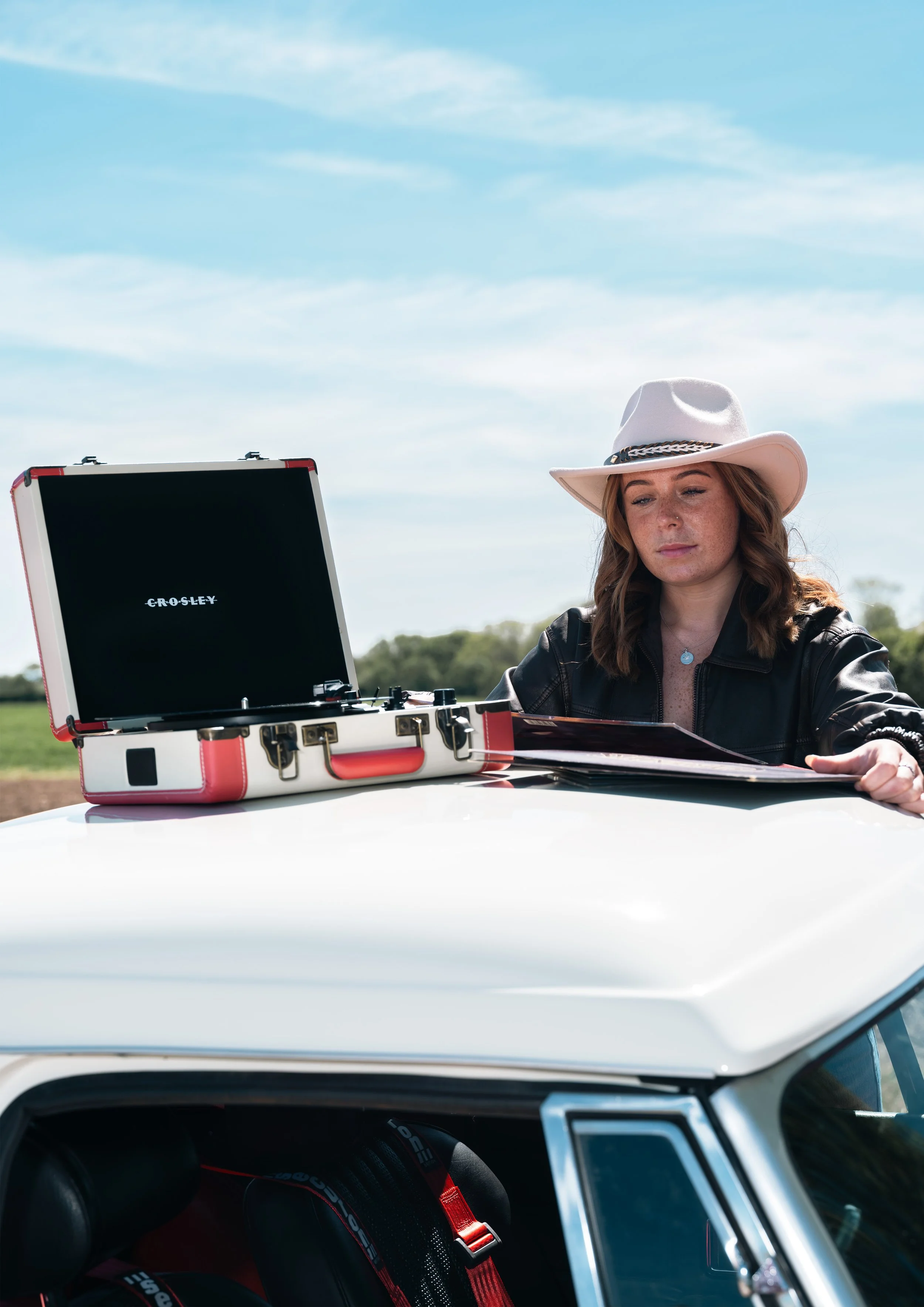 Woman in a white hat sitting at a table outside with a record player and papers, with blue sky and trees in the background.