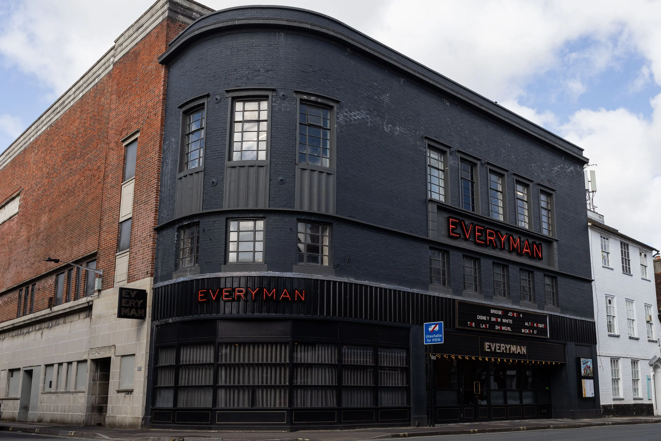The exterior of a building with a curved facade, painted black, and displaying the red neon sign for Everyman. The building has large, multi-pane windows and is situated on a city street under a partly cloudy sky.