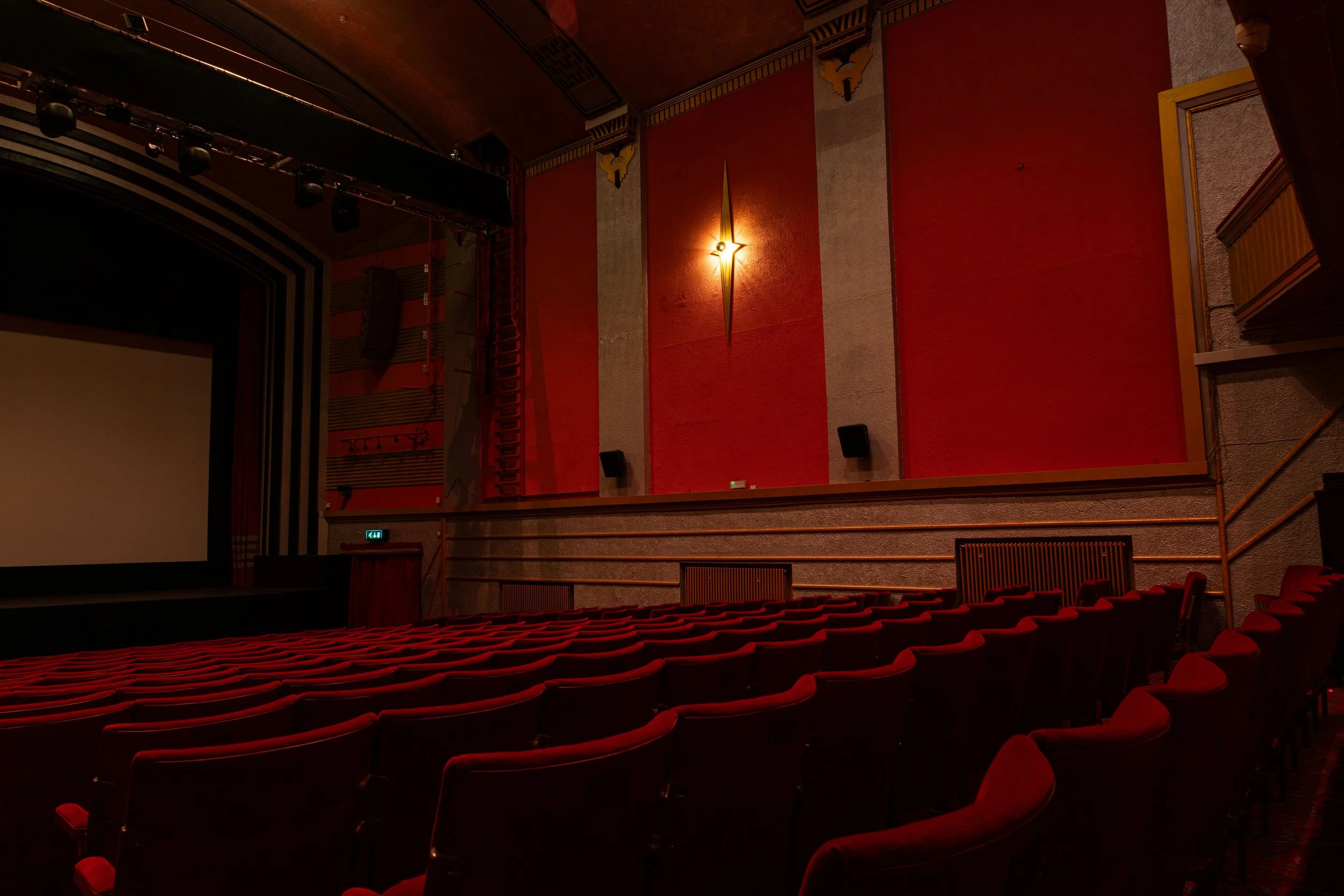 An empty movie theater with rows of red seats, a large projection screen, and a decorative feature on the wall with a light fixture.
