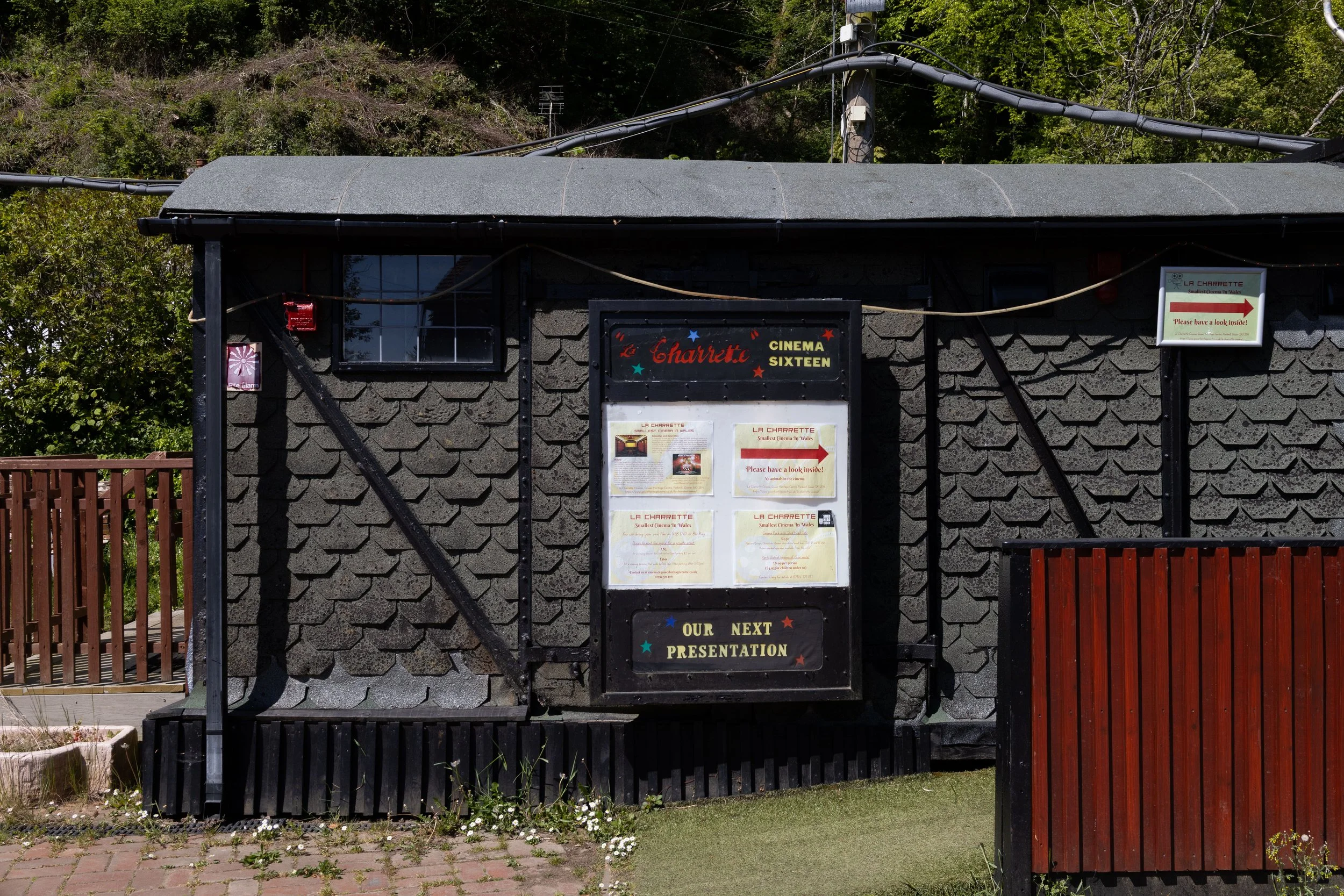 Small black building with a sign that reads 'La Charrette Cinema Sixteen' and posters for a movie called 'La Charrette'. There is a sign with an arrow and text welcoming visitors, and a small security alarm sign. The building has a wooden fence in fr