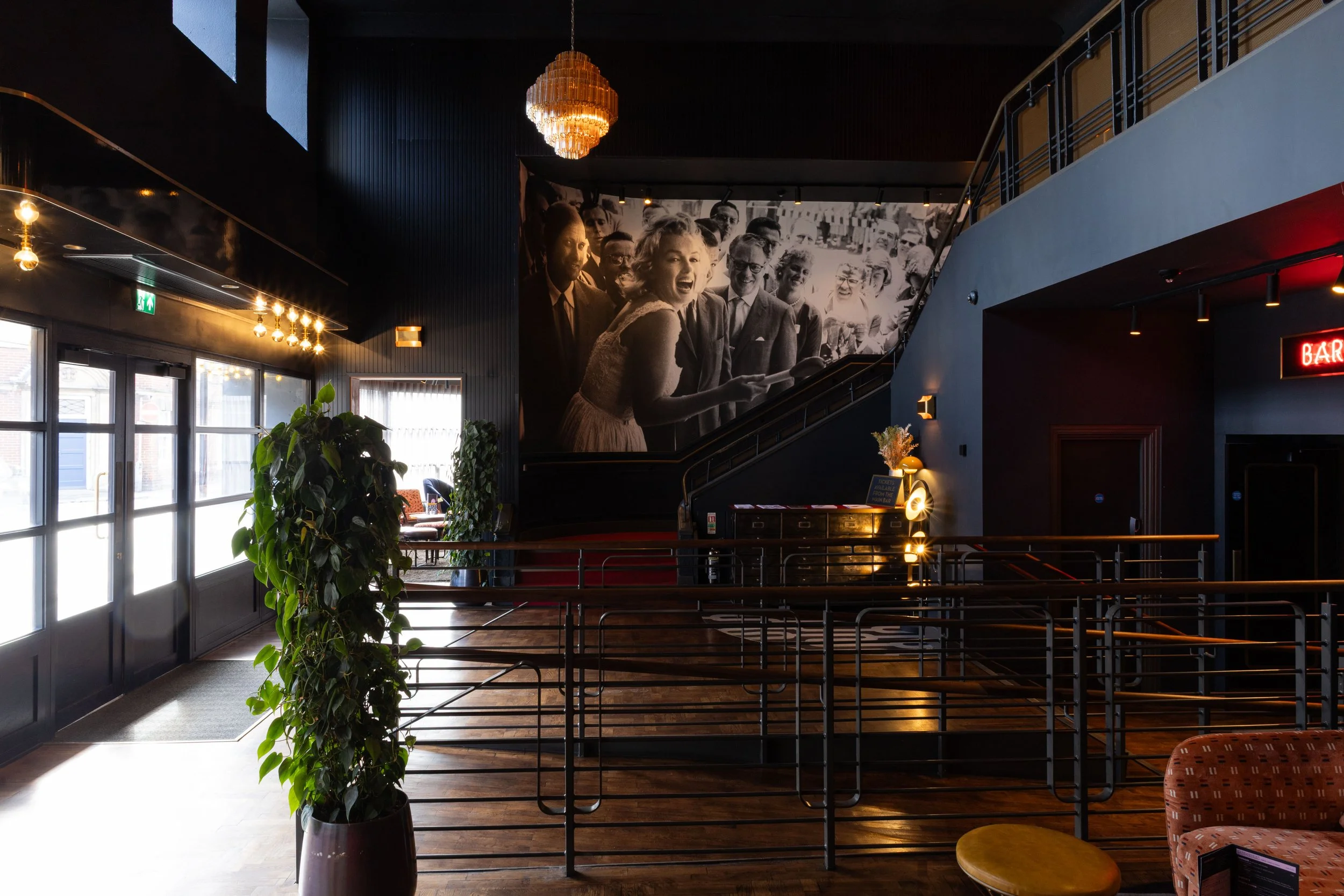 Interior of a modern building with dark walls, a black and white vintage-style photo of smiling people on the wall, a chandelier hanging from the ceiling, large windows letting in natural light, indoor plants, and a staircase with metal railings.