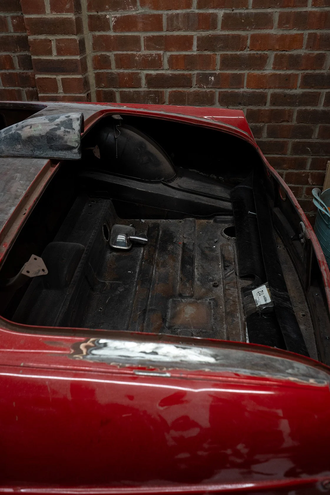 Interior of a vintage red car with no seats or interior components, showing a rusty and worn floor, and exposed black dashboard, against a brick wall background.