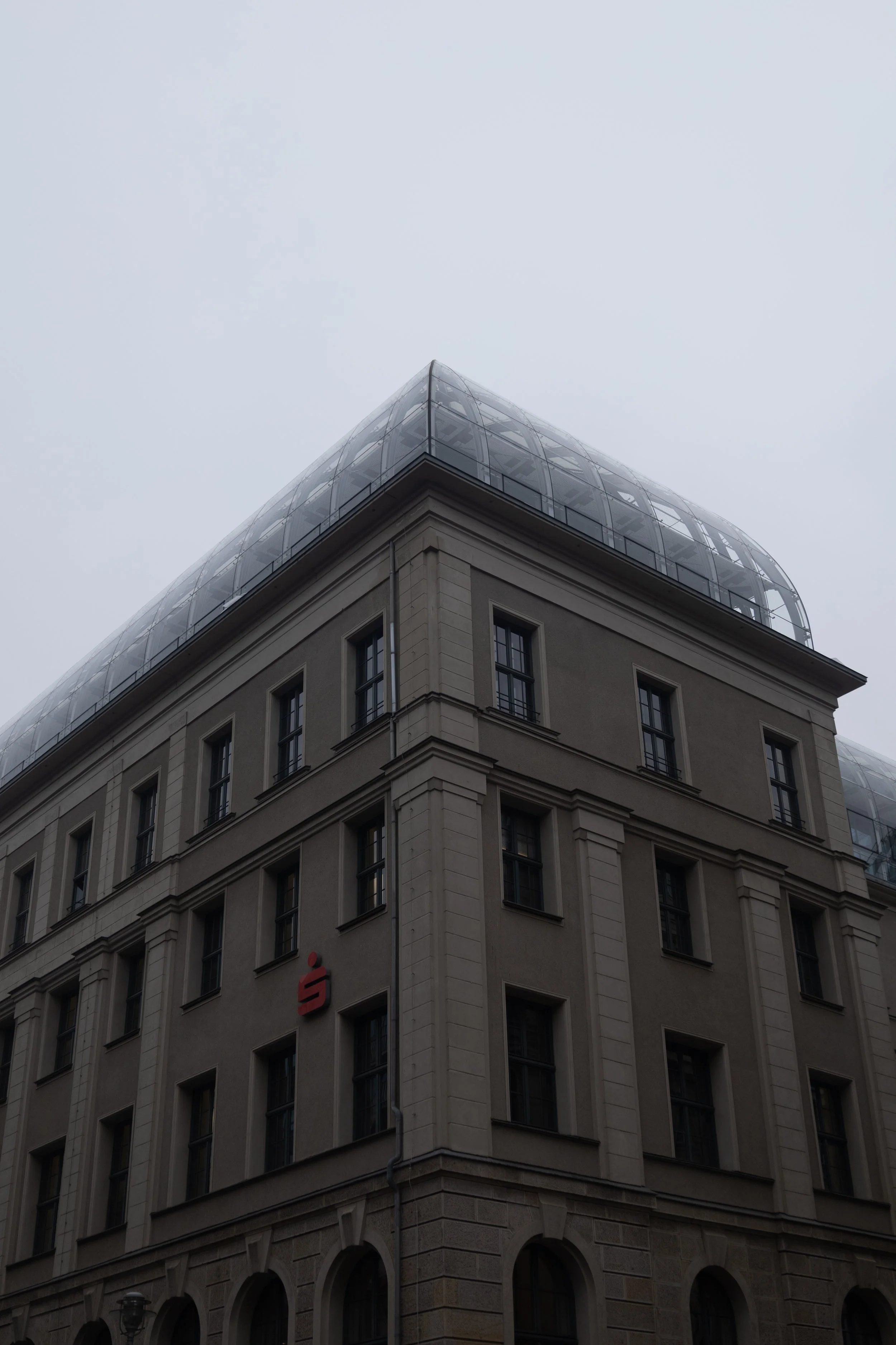 A corner of a tall building with multiple windows, a red logo, and a glass rooftop on a cloudy day.