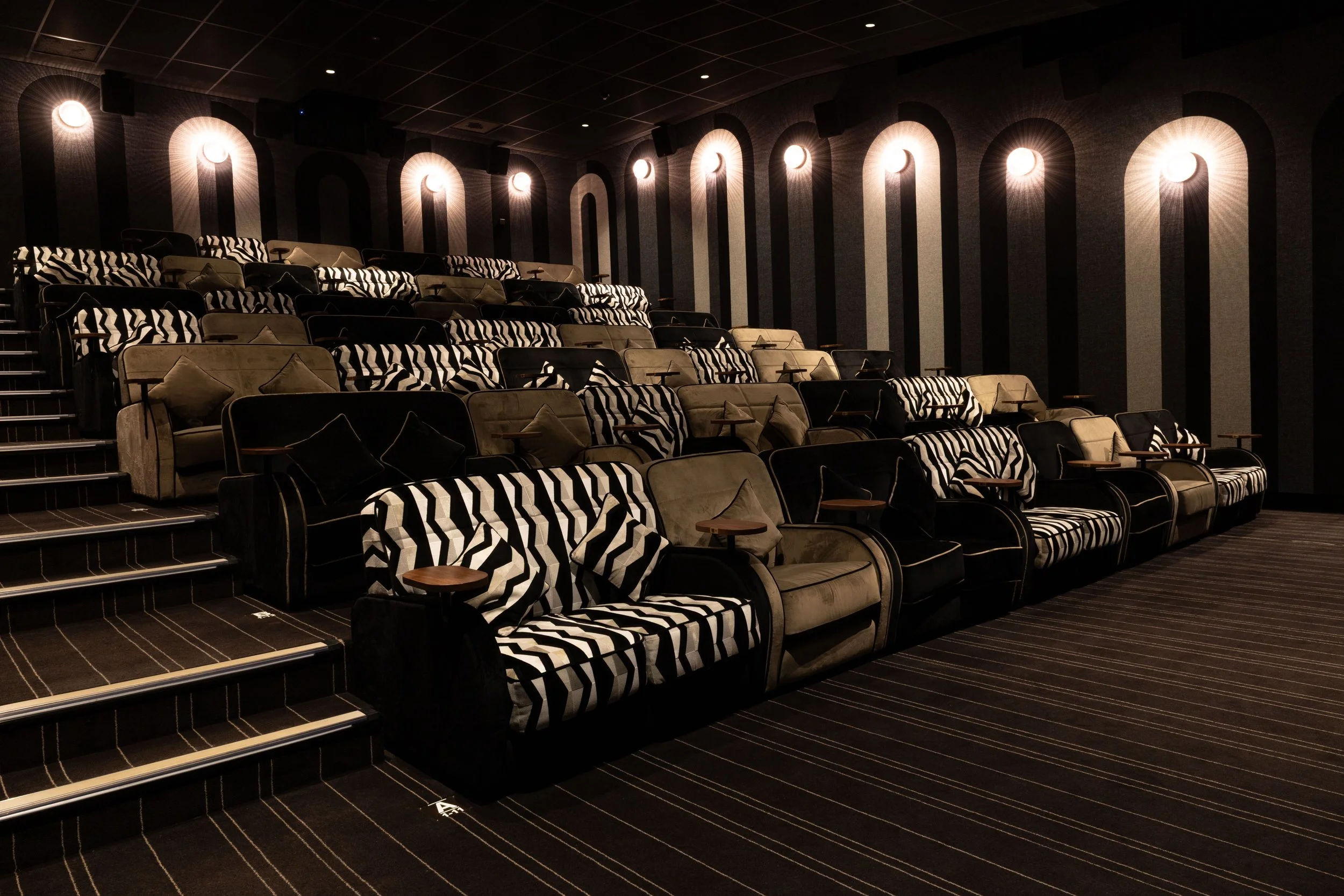 An empty movie theater with tiered rows of black and beige seats, some with black and white striped pillows. The wall behind the seats features vertical black and beige striped patterns illuminated by circular wall lights.