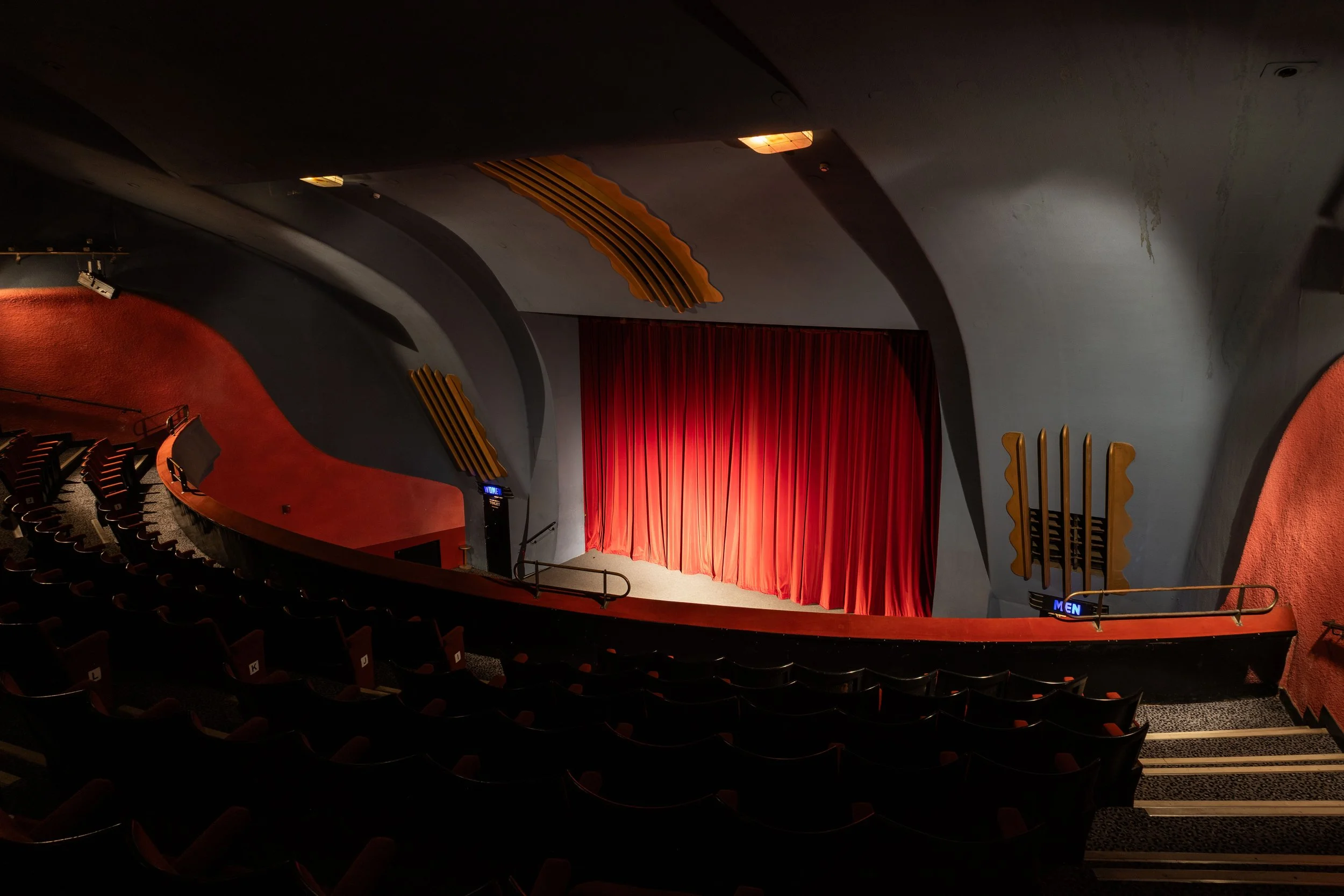 Inside a theater with a red curtain stage, curved balconies, and rows of empty seats, with some decorative yellow wooden wall elements and directional signs for men's and women's restrooms.