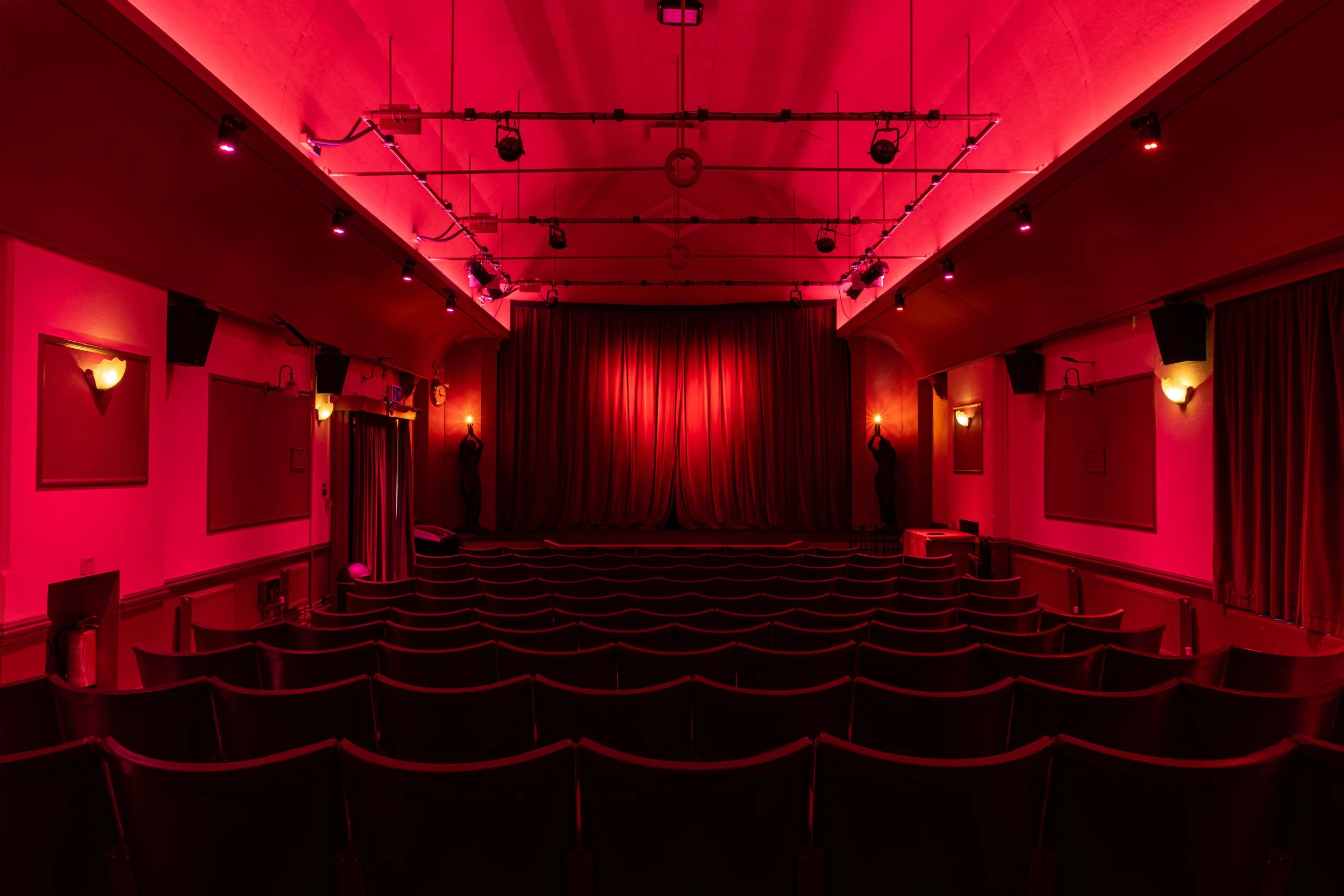 Empty theater with pink and red lighting, rows of seats, a stage with curtains, and stage lights hanging from the ceiling.