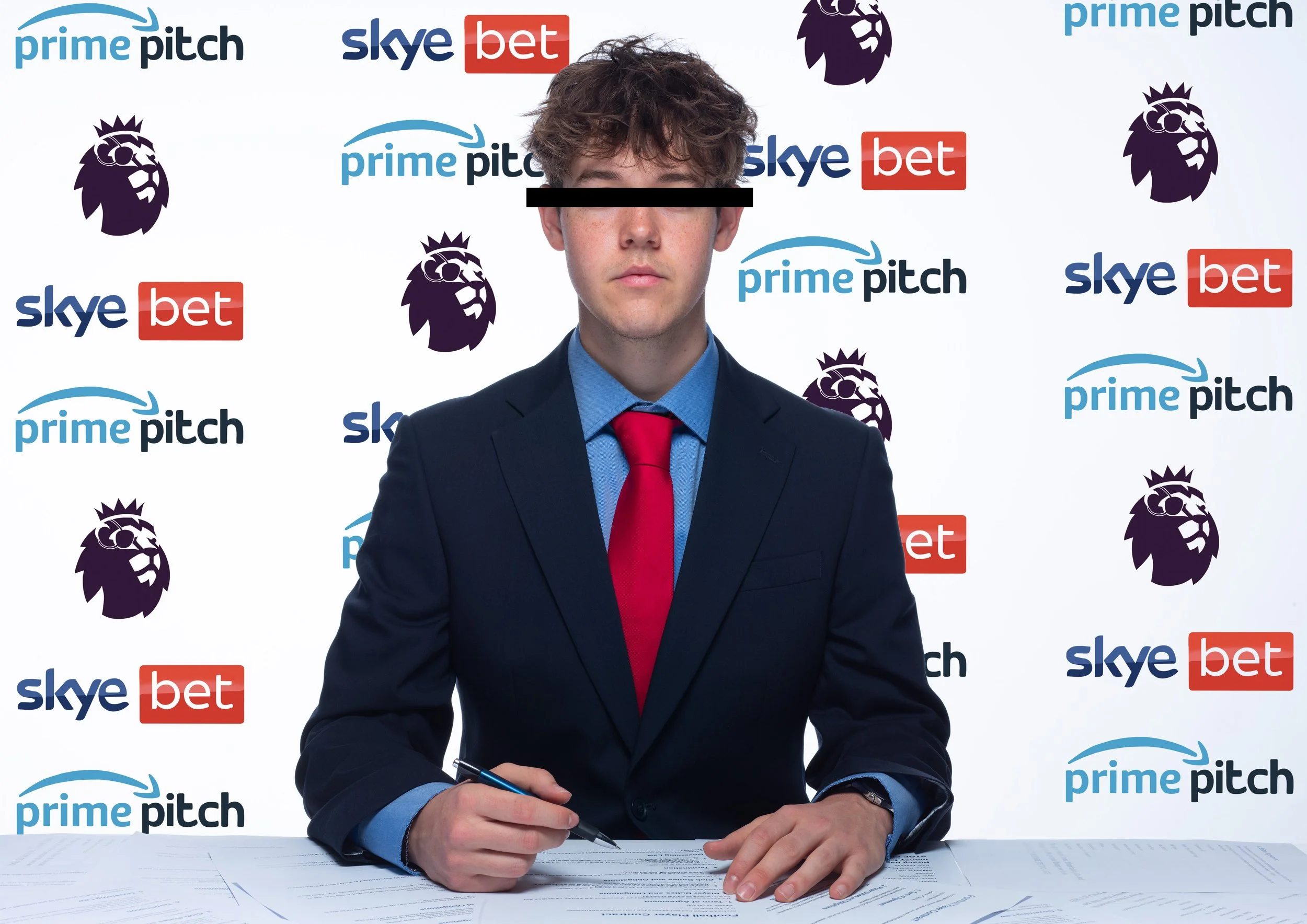 A young man in a suit and red tie is sitting at a table, holding a pen in his right hand, signing documents. The background features a white backdrop with logos of Prime Pitch, Sky Bet, and the Premier League lion logo repeated across it.