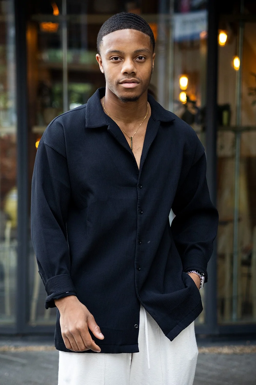 A young man with short hair wearing a black long-sleeve shirt and light-colored pants stands outside in front of a glass building.