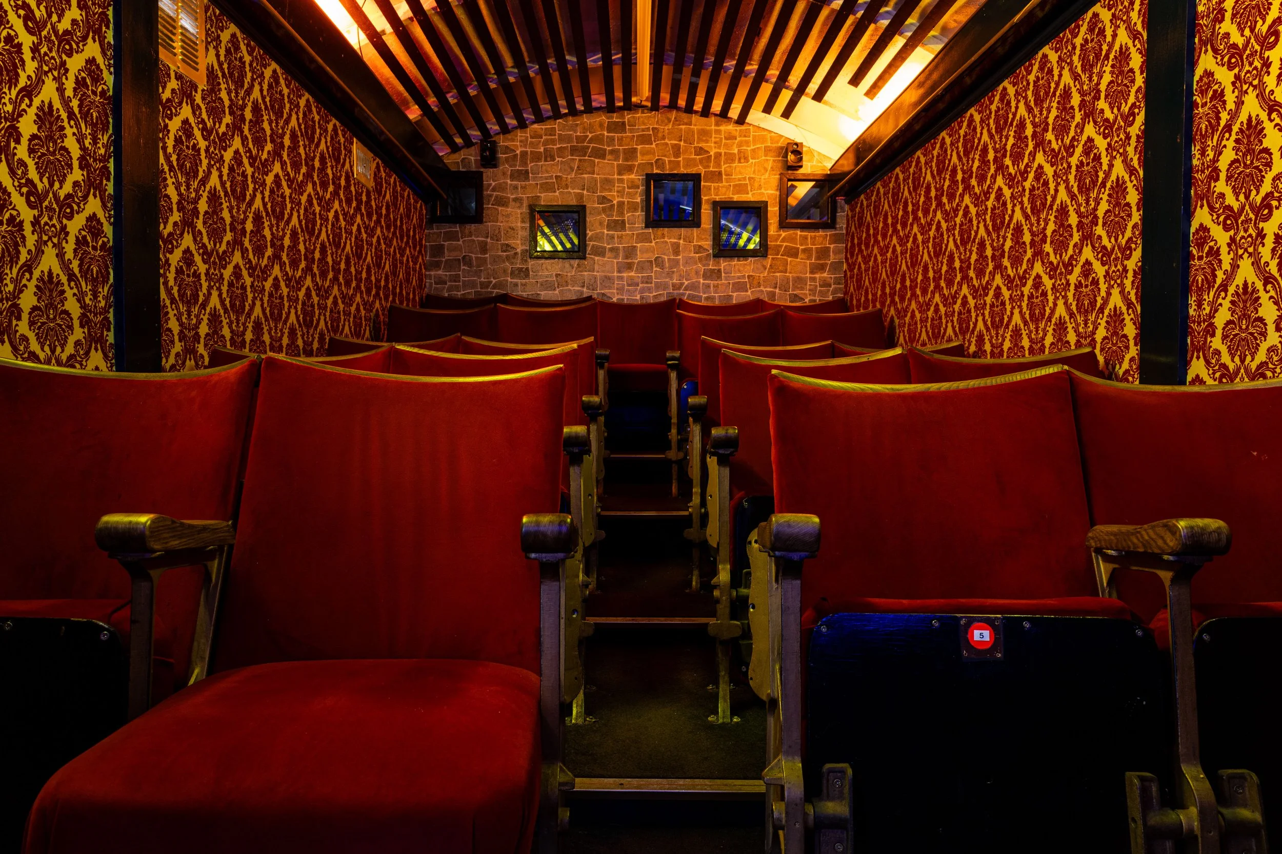 Empty red theater seats facing a stone wall with four small flat-screen monitors and some black frames hanging on the wall. The ceiling features wooden beams with colorful lighting.