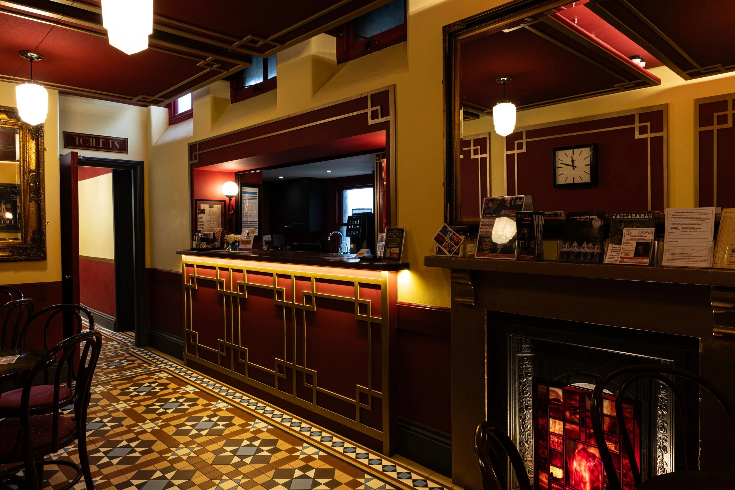 Interior of a restaurant or cafe with a decorated counter, patterned tile floor, and vintage-style lighting fixtures. There is a sign indicating the bathroom and a clock on the wall.