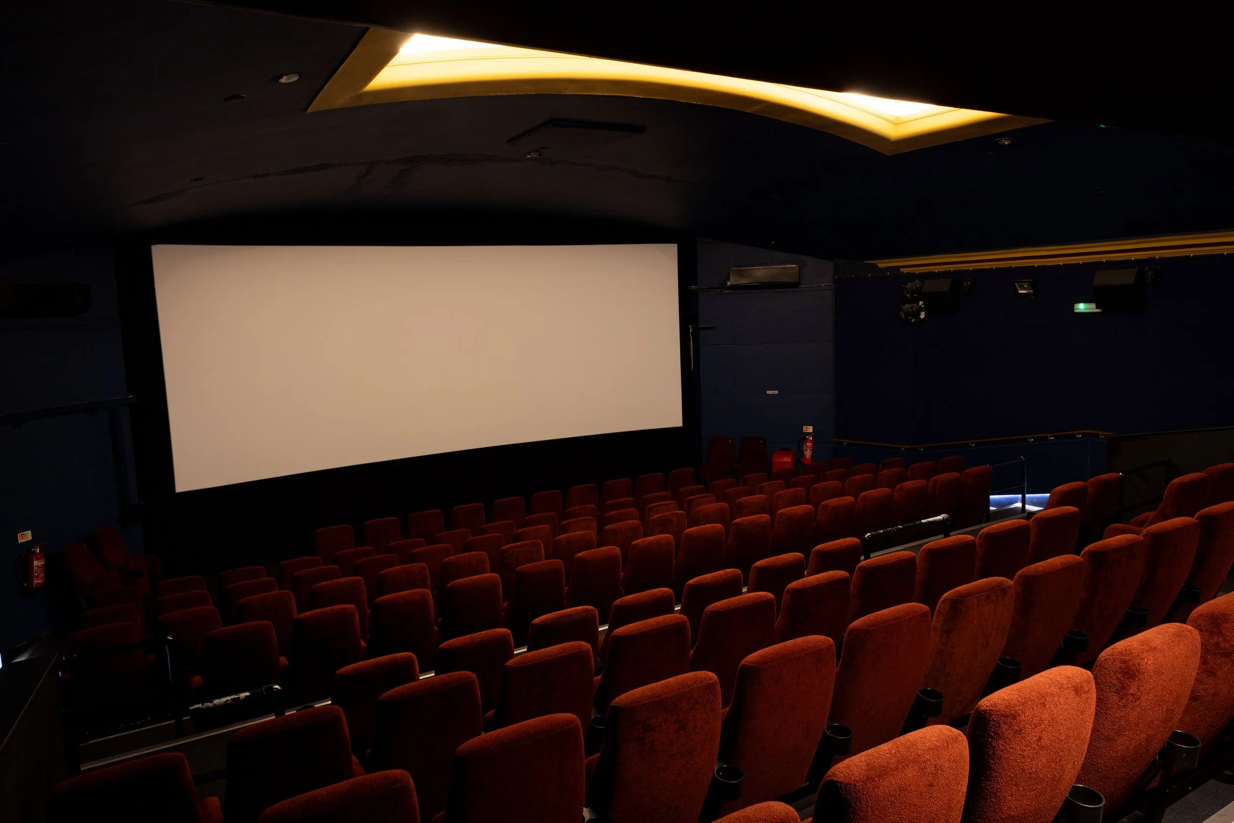 Interior of a movie theater with rows of red velvet seats and a large blank projection screen on the front wall.