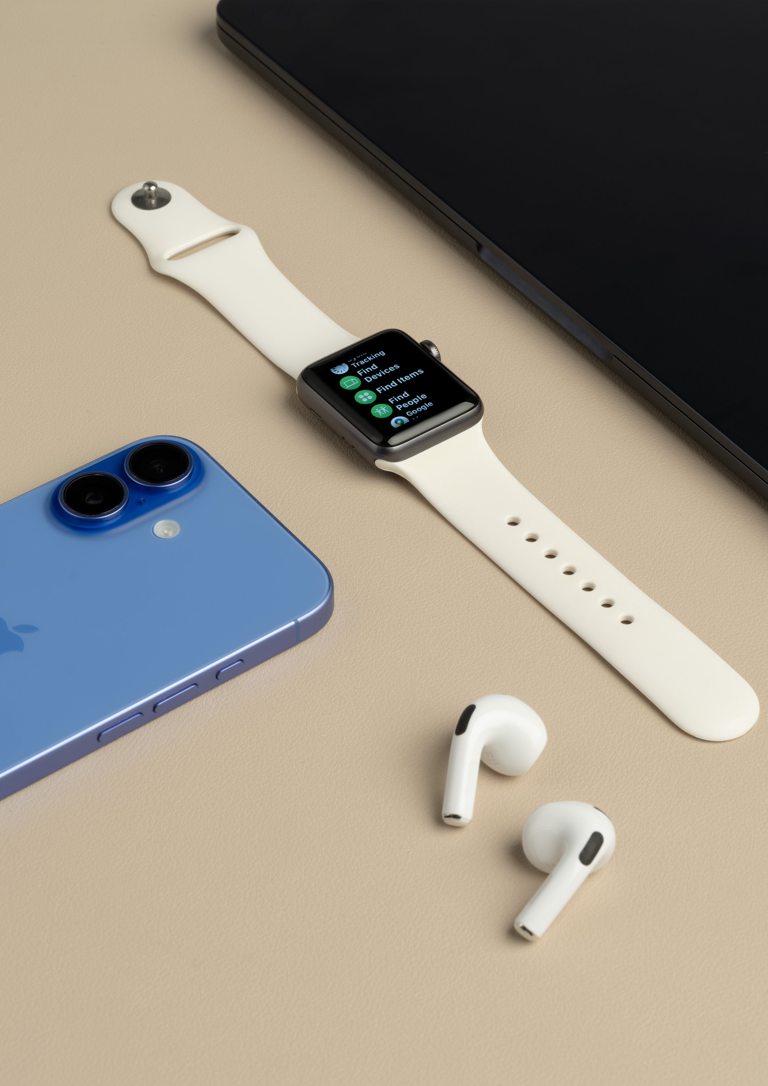 An Apple Watch with a white band and a screen displaying the 'Find' app, an iPhone with blue back and triple camera, wireless earbuds, and a dark laptop on a beige surface.