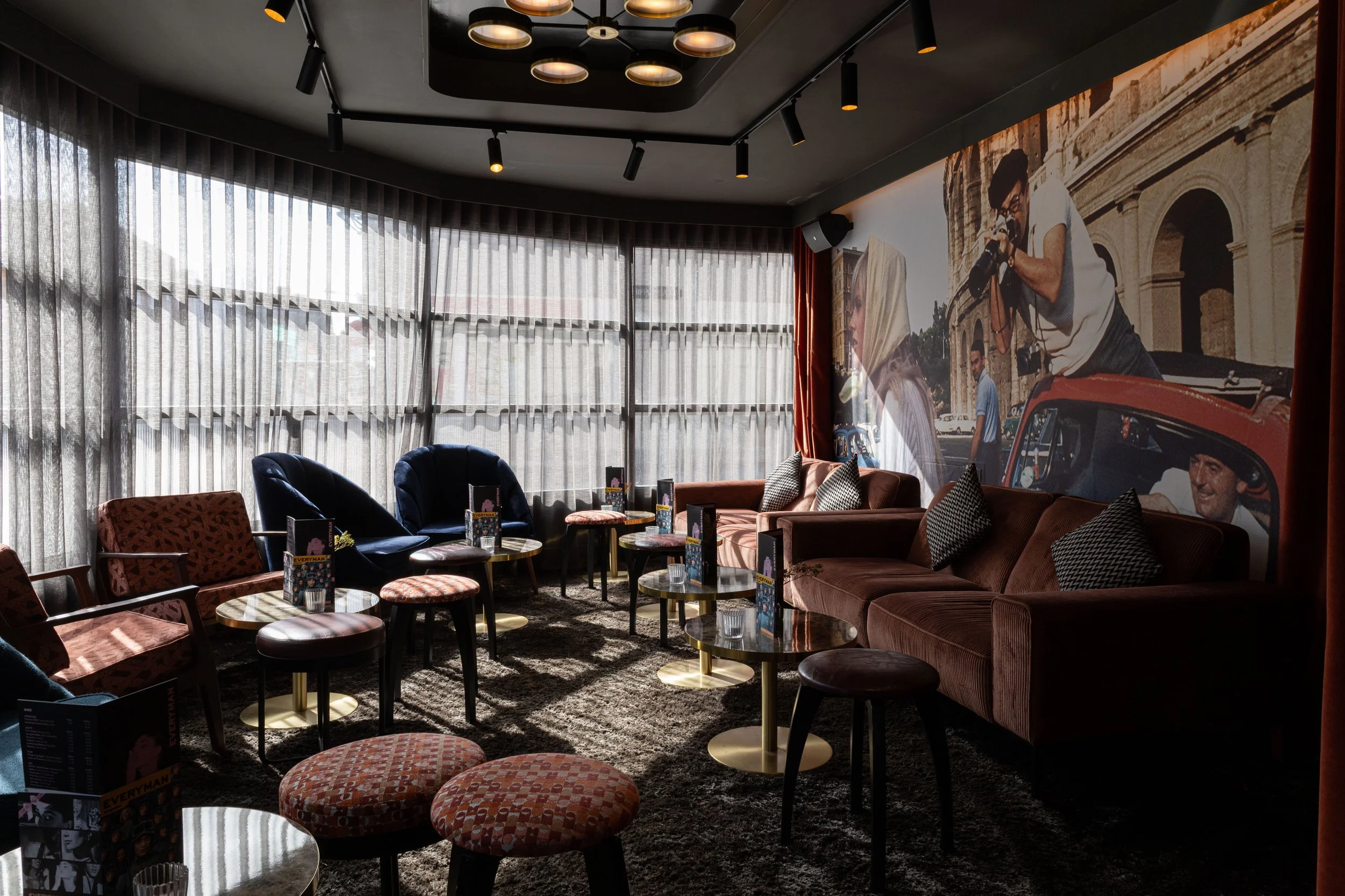 Interior of a lounge or cafe with a large mural on the wall depicting people taking photos in front of historic landmarks, with curved windows and sheer curtains.