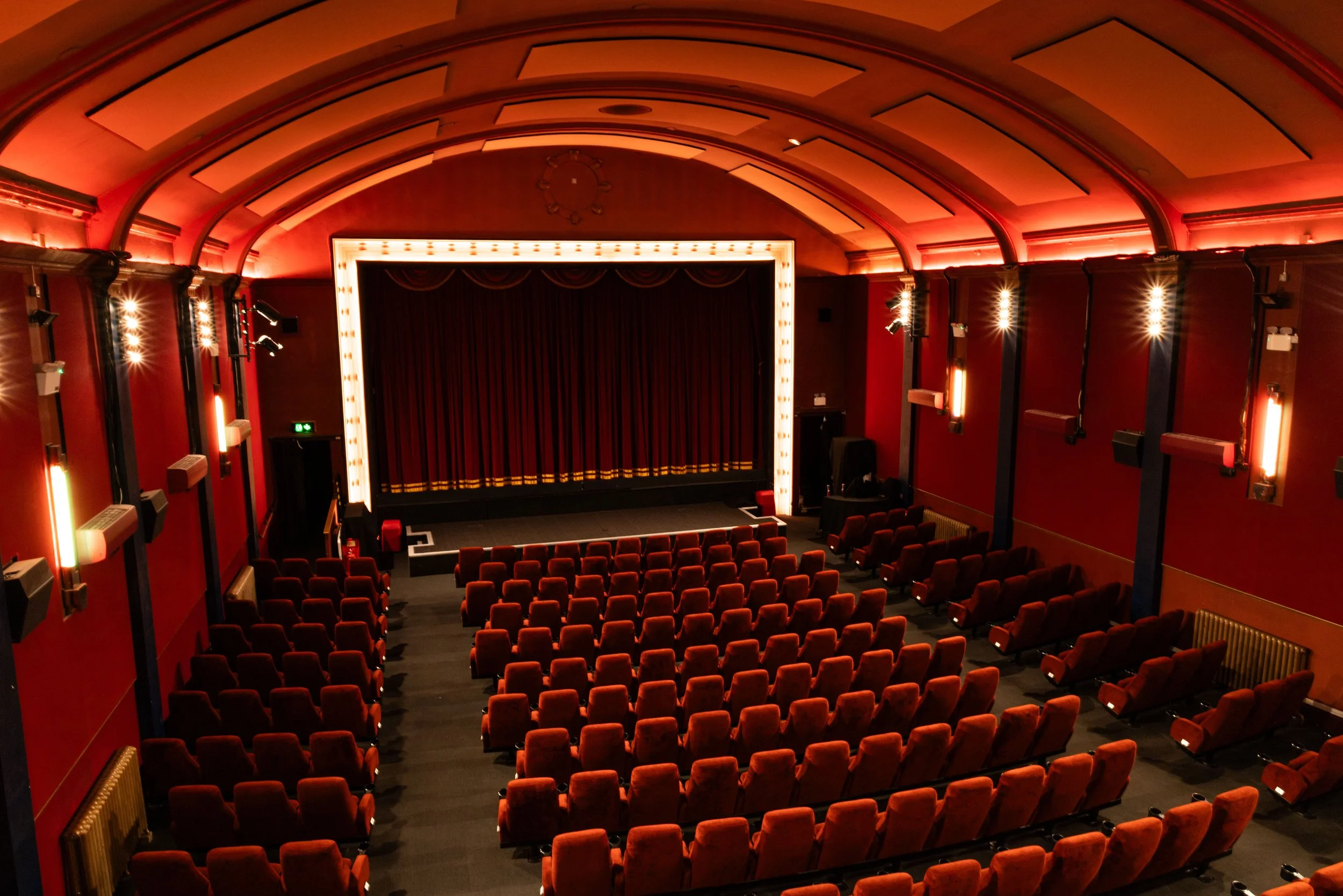 The interior of a theater with red walls, a stage with a curtain, red seats, and decorative lighting along the walls and ceiling.
