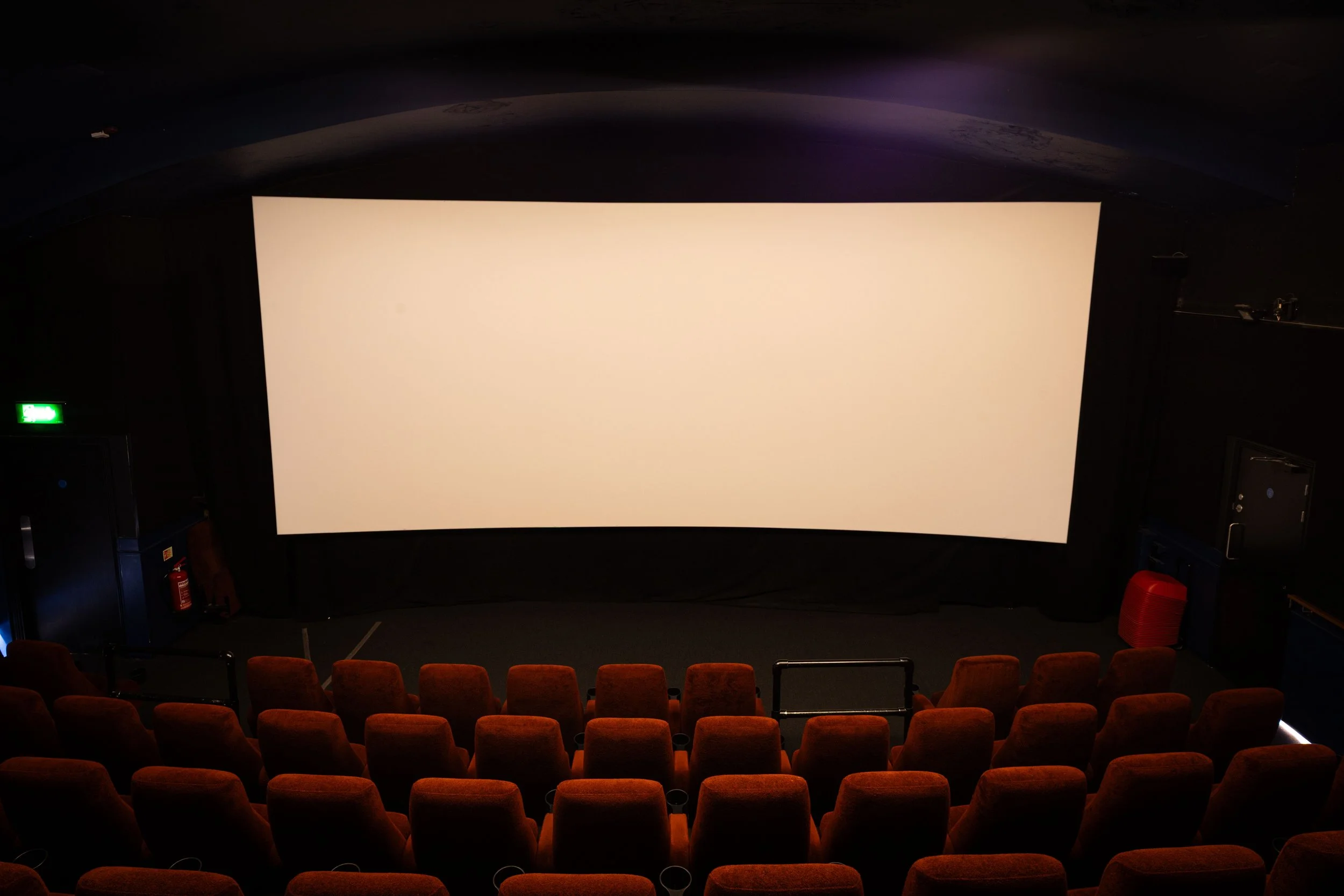 A movie theater auditorium with red seats, a large blank projection screen at the front, and dim lighting.