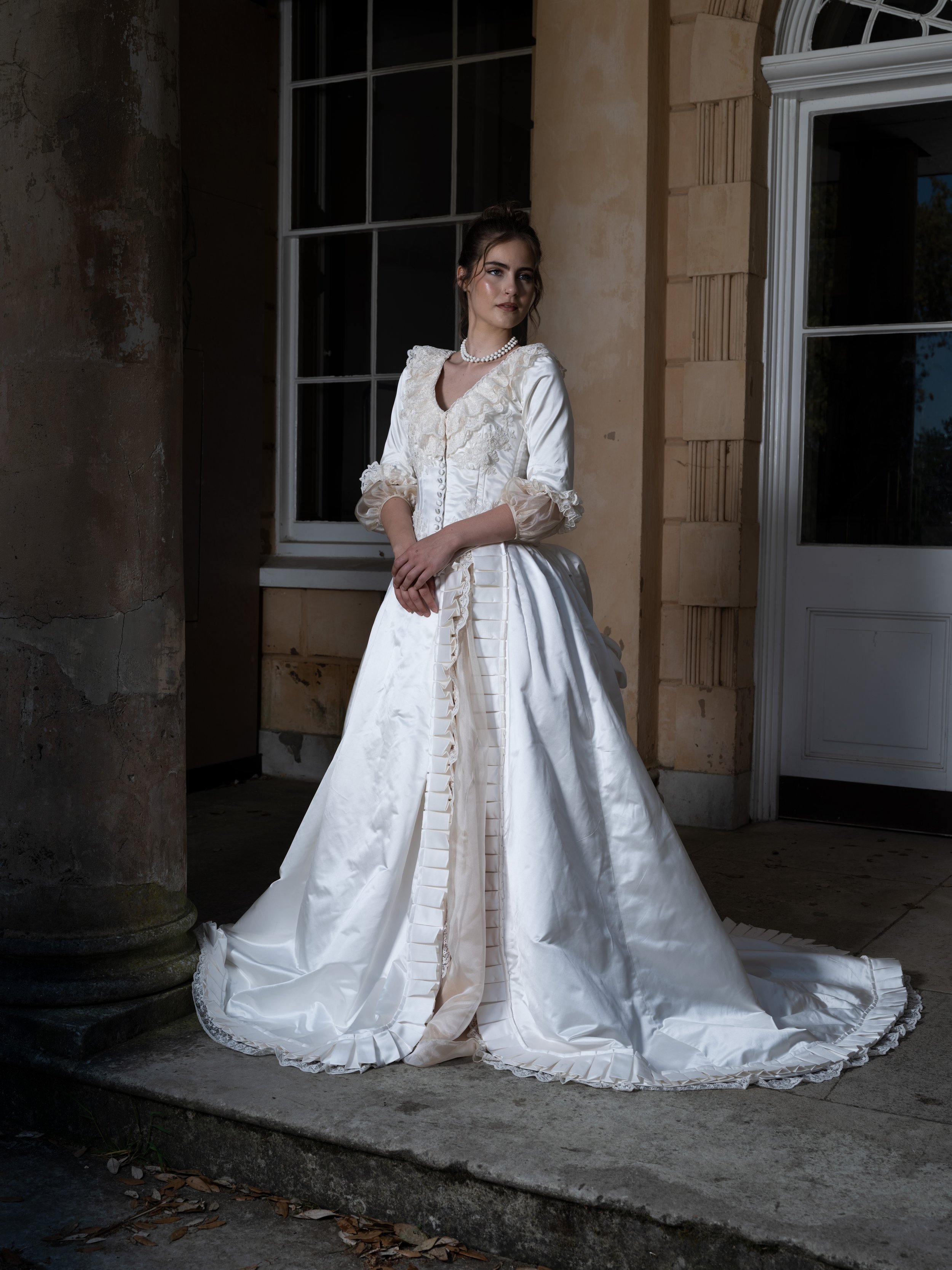 A woman in a vintage white wedding gown with lace and puffed sleeves poses in front of a stone building with large windows, standing on a stone step.