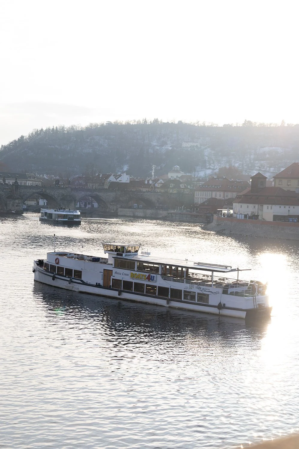 A river cruise boat on a calm river with a backdrop of a cityscape and hills covered in snow, under bright sunlight.