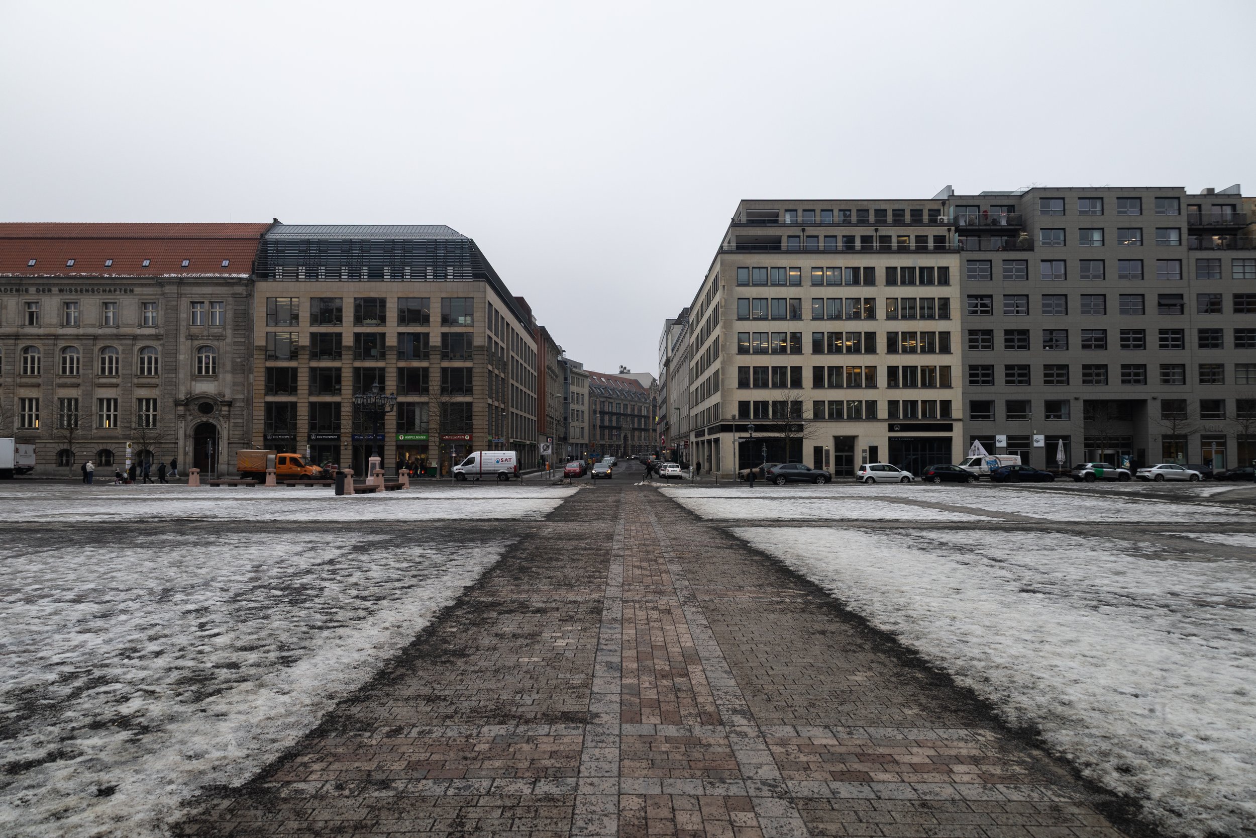 City street with modern buildings, overcast sky, and patches of snow and ice on the ground, with parked cars and pedestrians.