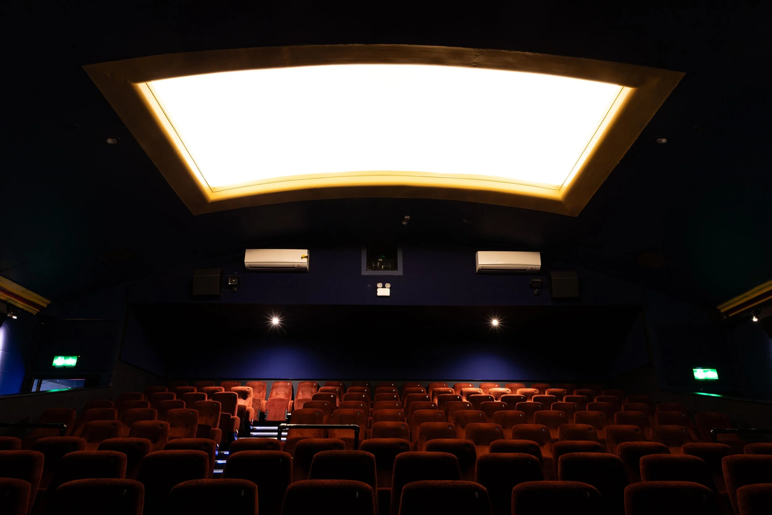 Empty movie theater auditorium with rows of red seats, a large illuminated ceiling light, and dark walls.