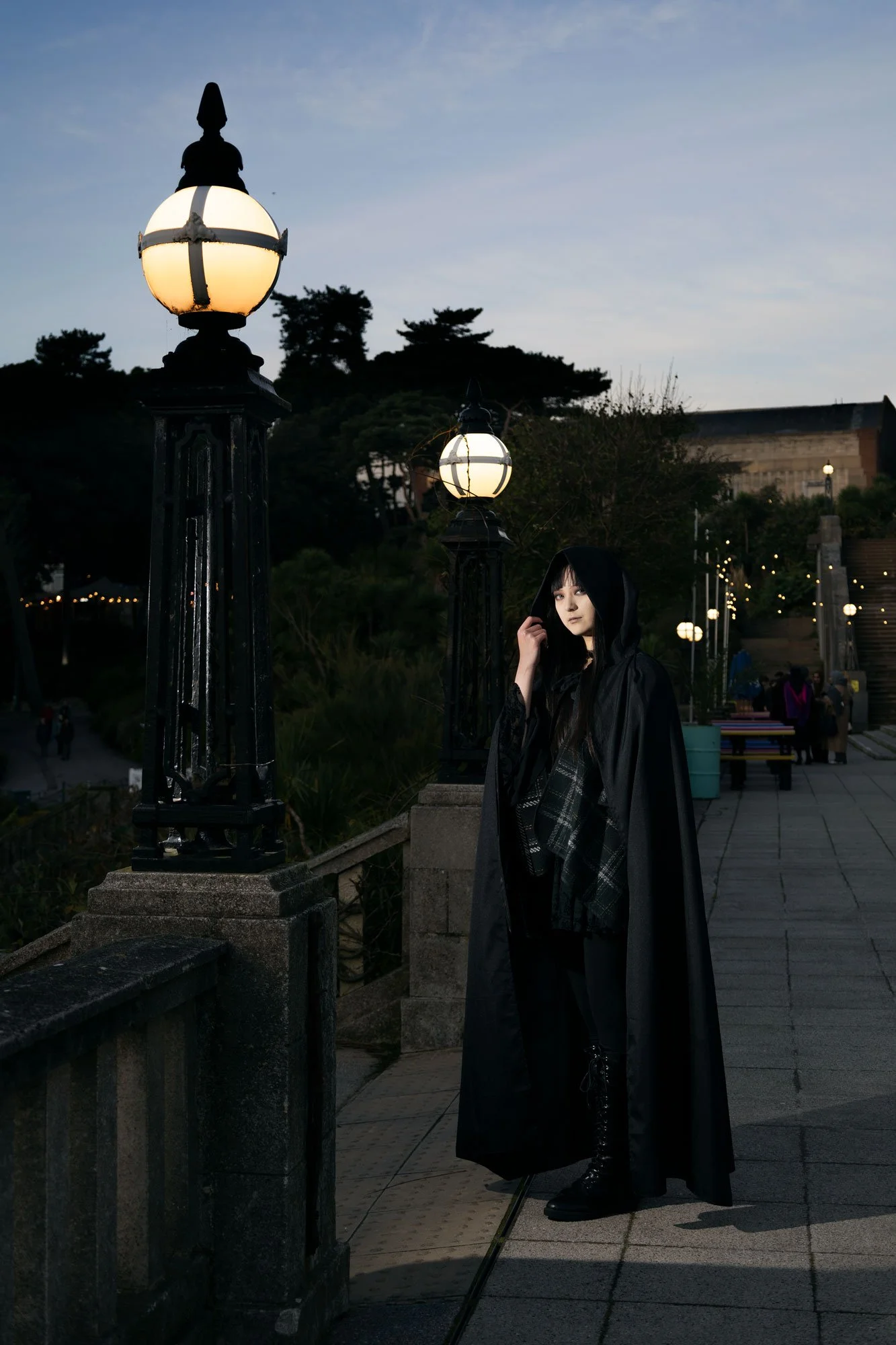 A woman dressed in goth attire wearing a long black cape with a hoodie, standing outdoors during dusk near vintage-style street lamps, with trees, buildings, and a staircase in the background.