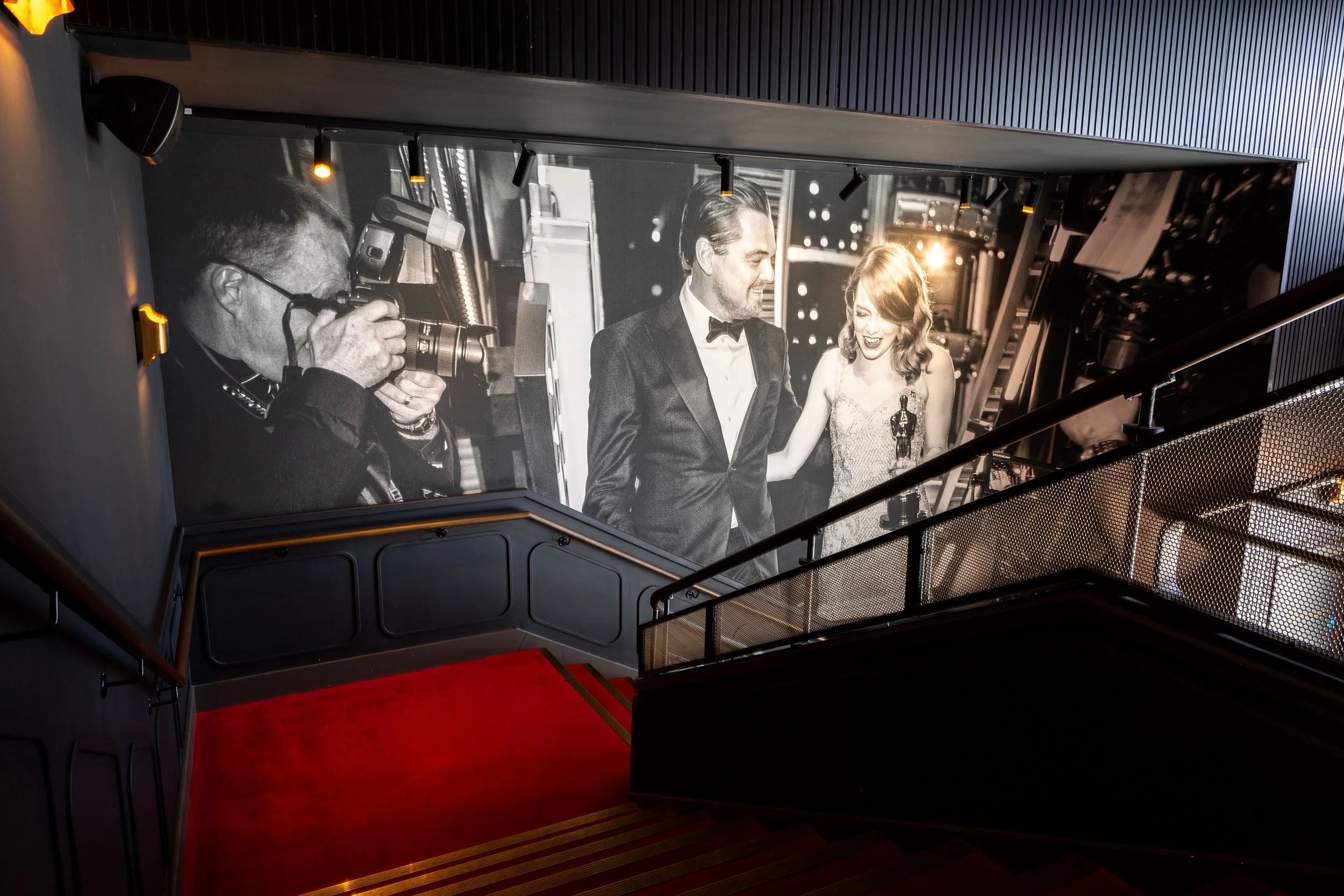 Black and white photo on a large screen showing a man and woman laughing at an event, with a photographer taking a picture of them.