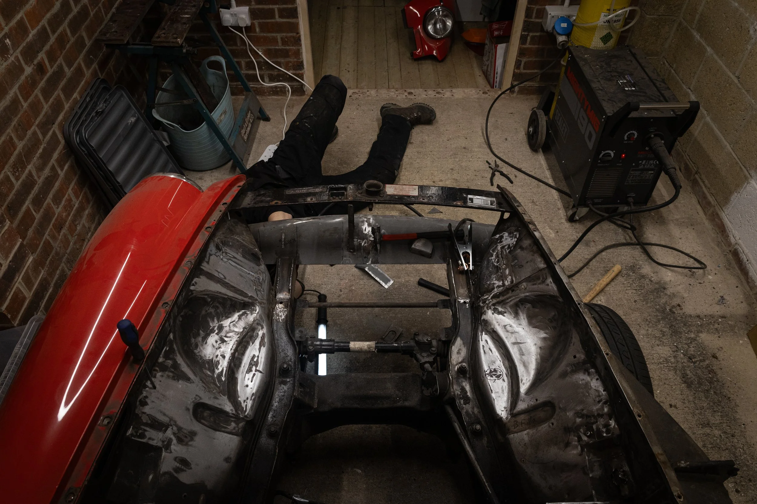 A person working on a car in a garage, lying on the floor with legs spread out, welding equipment nearby, and tools scattered around.