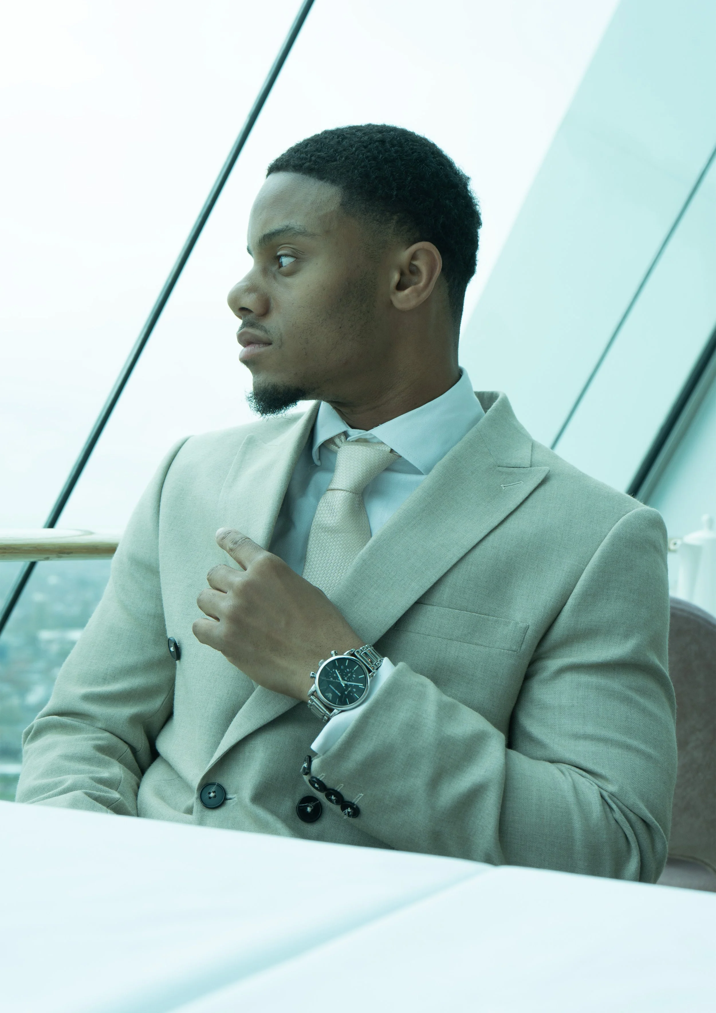 A man wearing a beige suit with a tie, looking thoughtfully to the side with his hand over his chest, in front of large windows with a city view.
