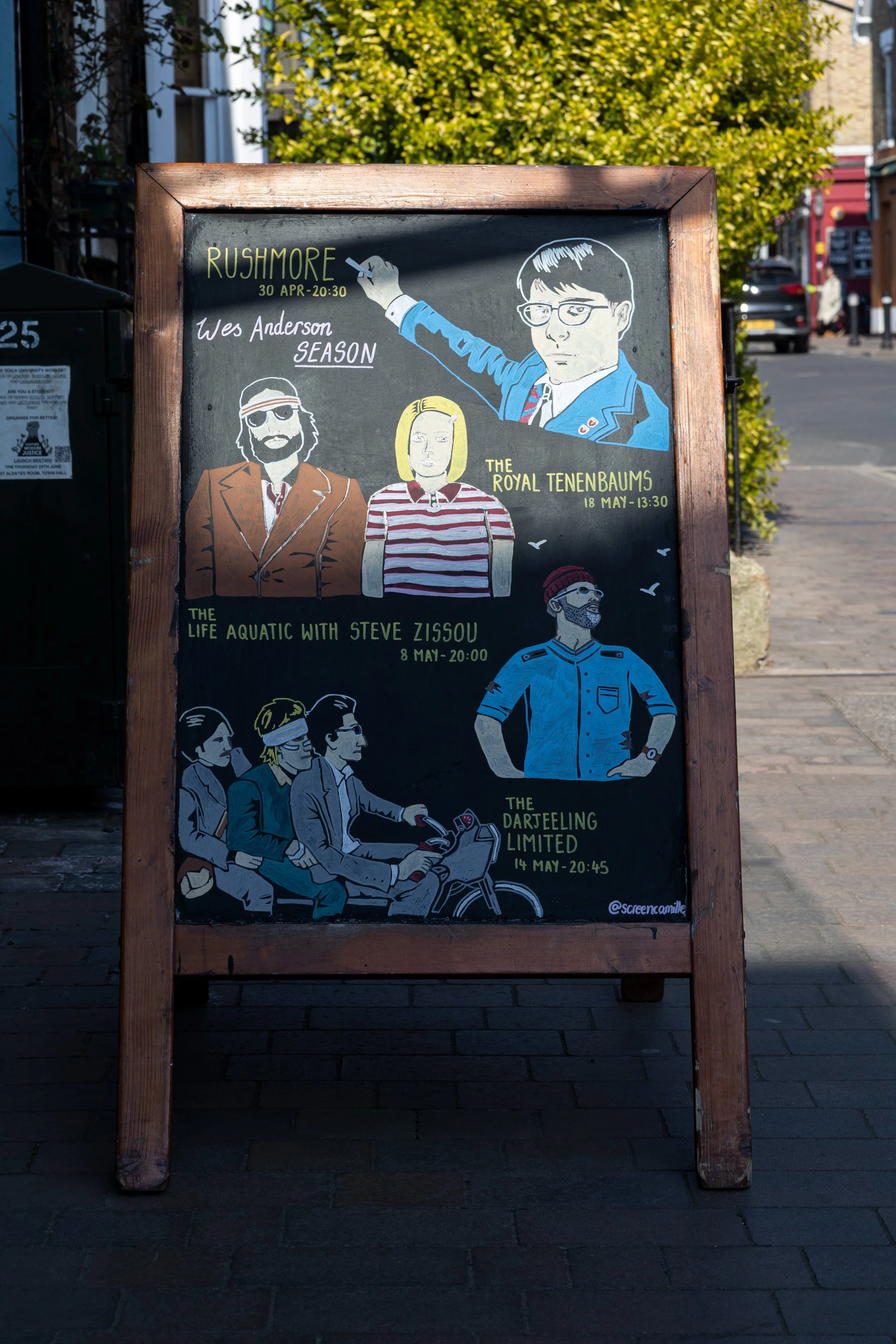 A chalkboard sign on a sidewalk displays illustrated poster advertisements for upcoming performances, including bands and comedians, with details about dates and times. The background shows a street scene with a tree and buildings.