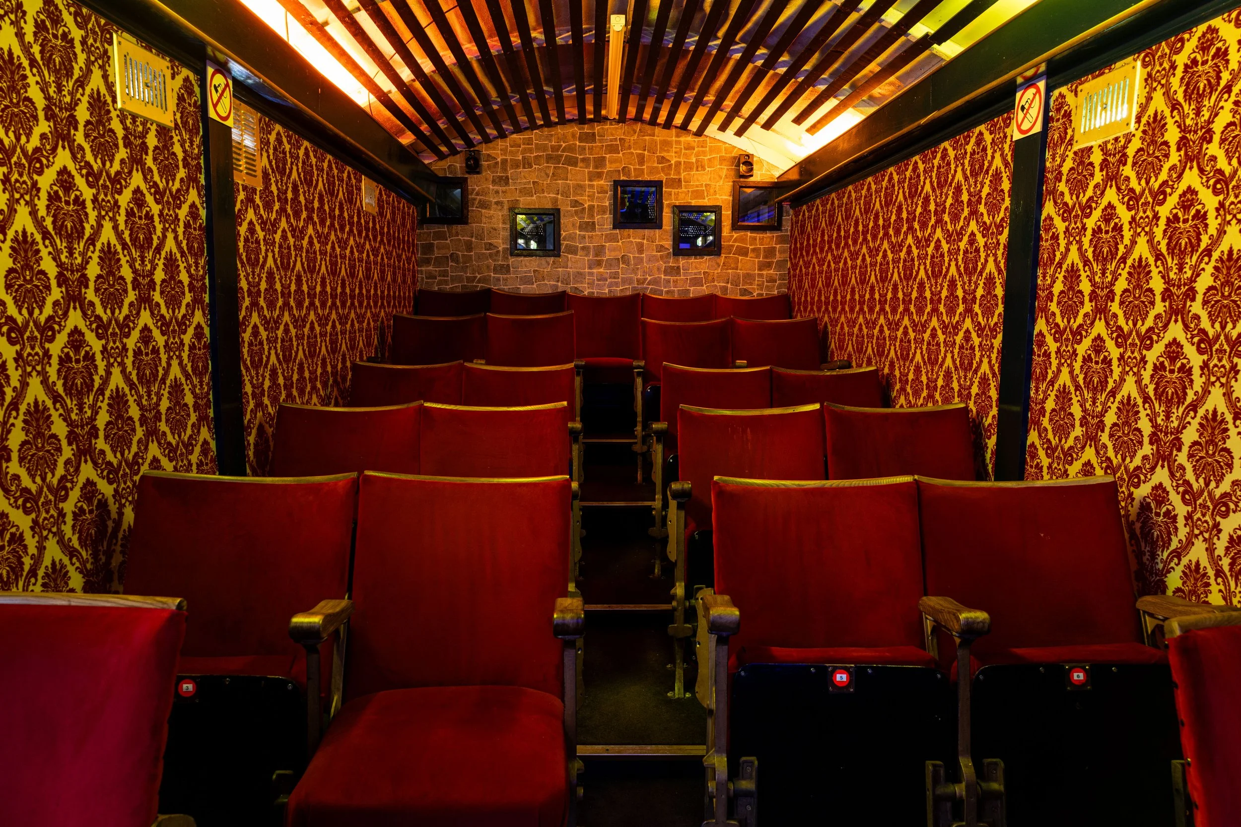Empty vintage theater or auditorium with red velvet chairs, decorative yellow and maroon wallpaper, brick wall with four small framed windows, wooden ceiling beams, and overhead lighting.