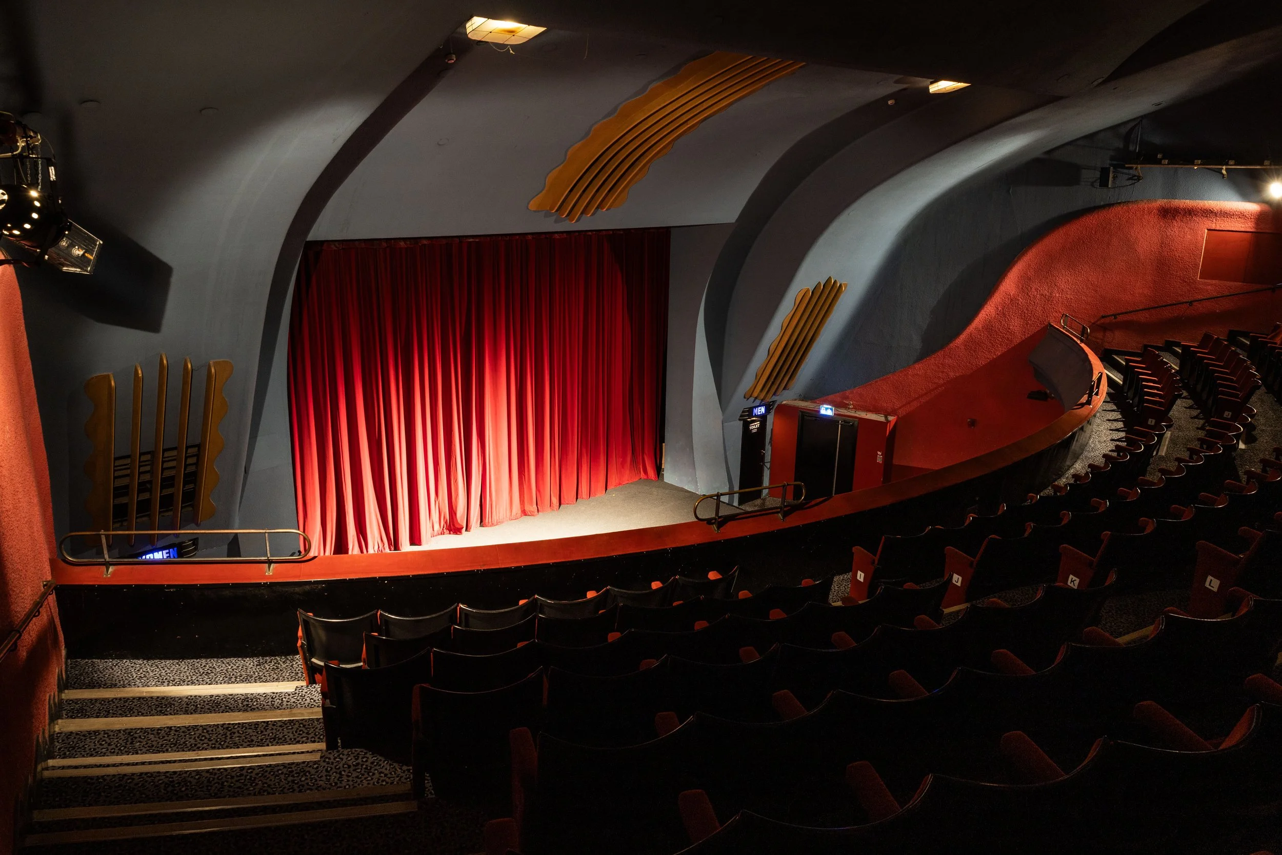 An empty movie theater with red velvet curtains, black seats with red accents, and ornate gold decorations on the walls and ceiling.