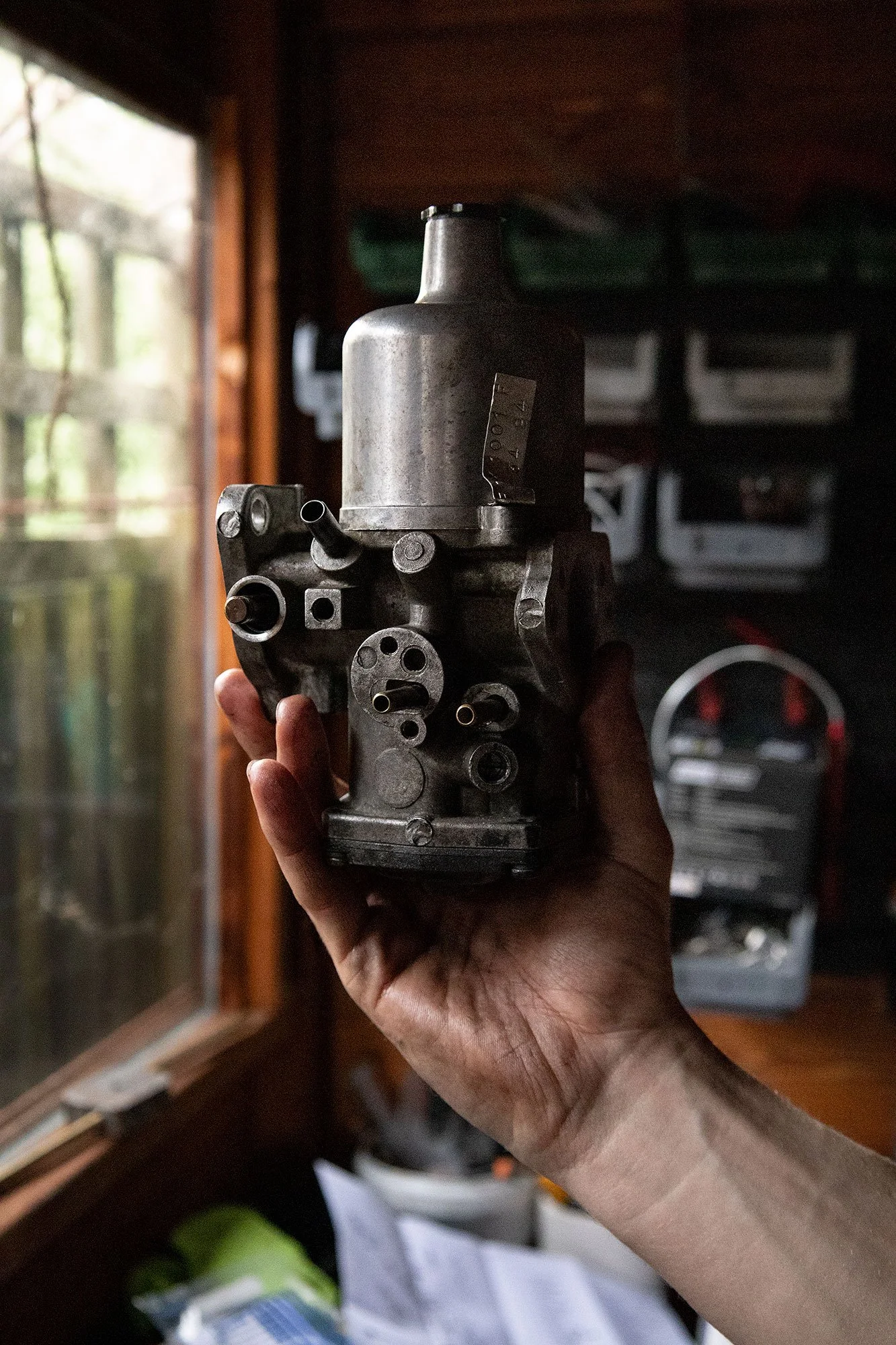A hand holding a metallic mechanical part or component near a window with blurred background.