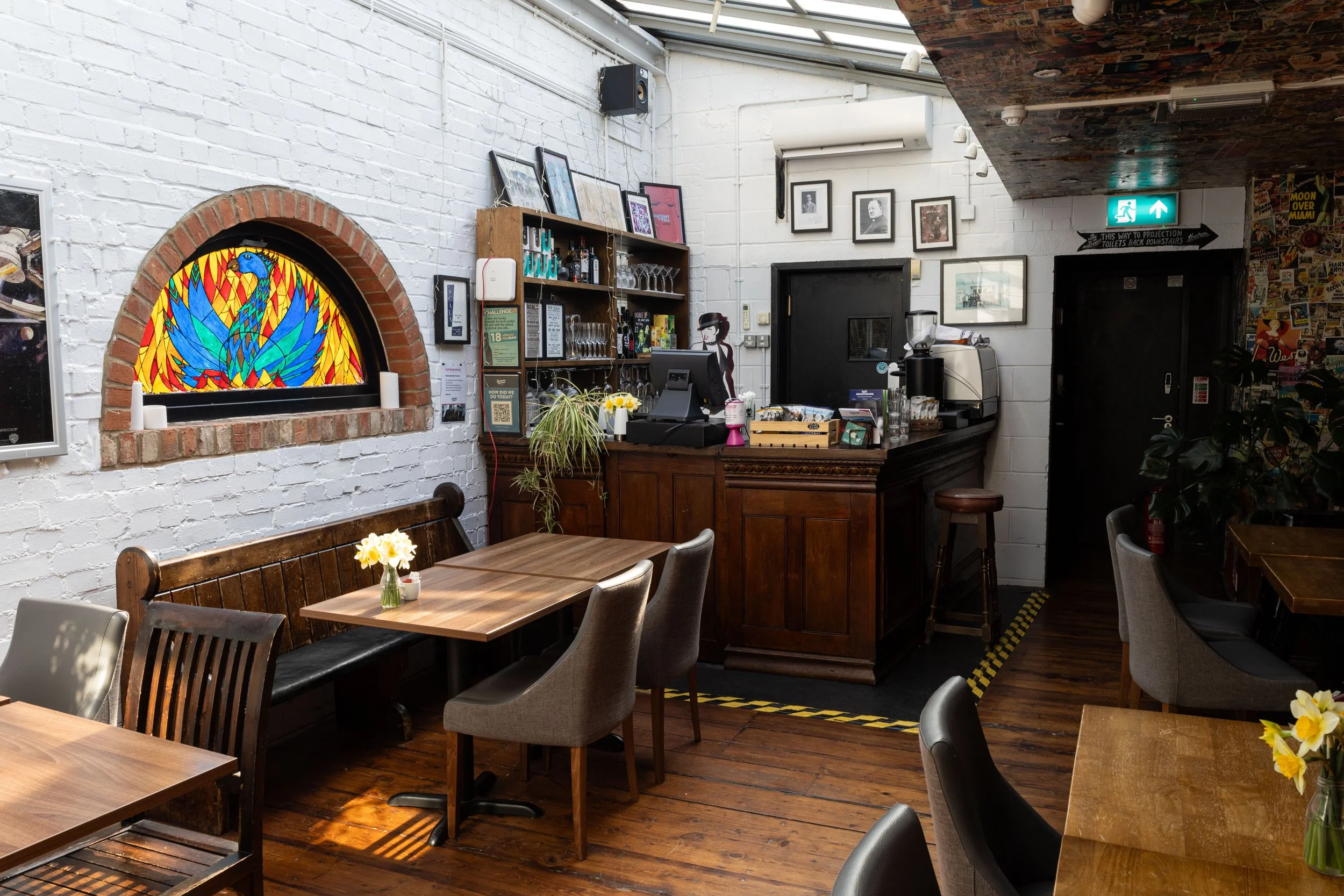 Interior of a cozy cafe with wooden tables and chairs, white painted brick walls, photo frames, a stained glass window of a colorful bird, and a bar area with glasses and bottles, illuminated by natural light and ceiling lights.