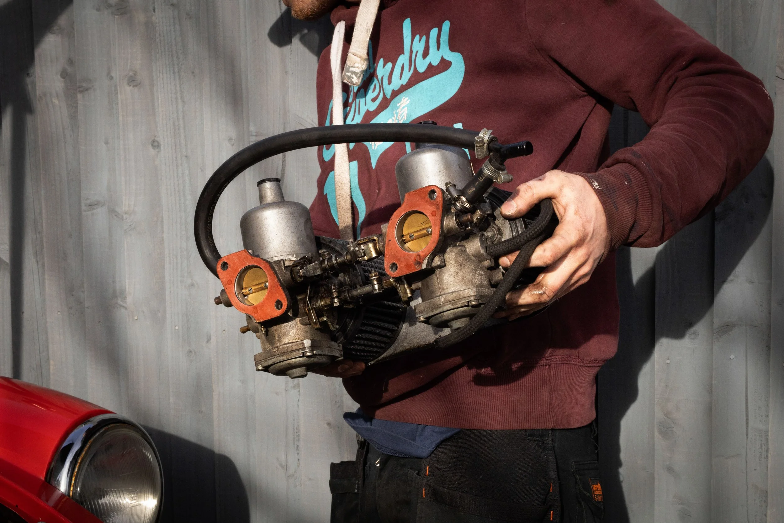 A person wearing a maroon hoodie with blue text and black pants is holding a disassembled carburetor or small engine part in front of a gray wooden wall. The person's face is not visible.