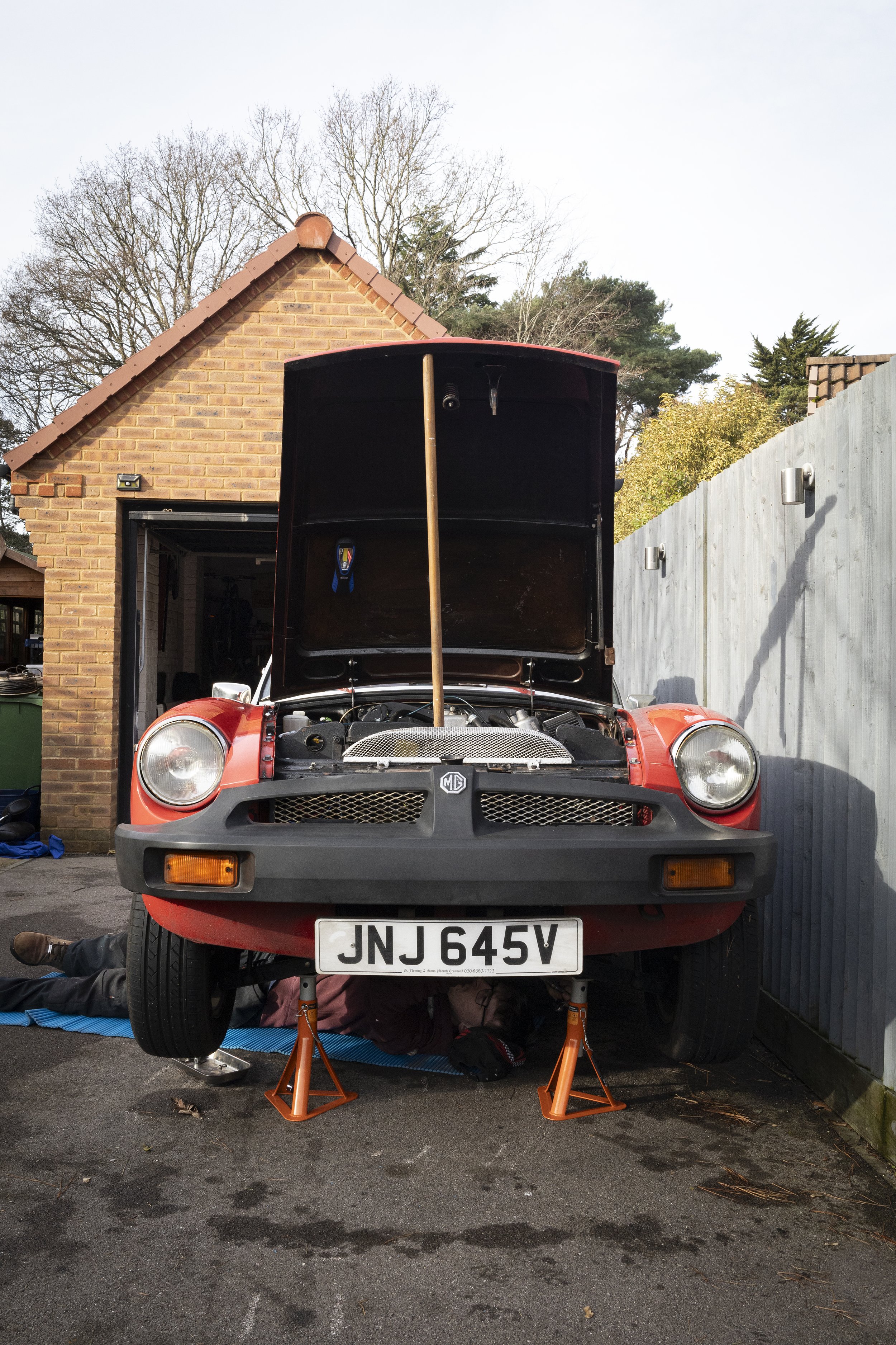 A red sports car with its hood open, parked in a driveway. The car is supported by orange jack stands and appears to be undergoing maintenance. There is a person working underneath the car, with only their legs visible. The background shows a brick h