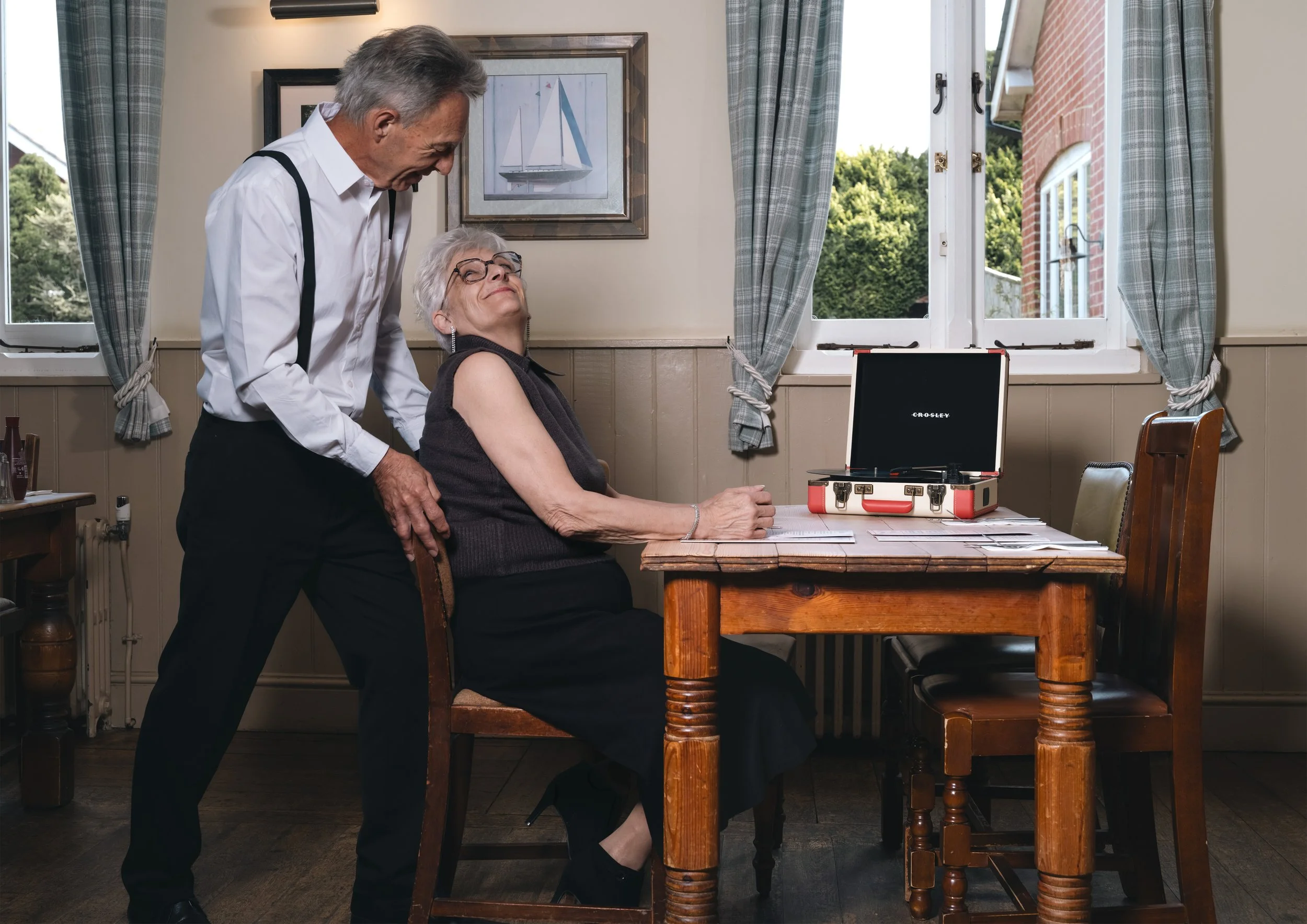 An older man and woman are in a cozy kitchen. The man is leaning over the woman who is sitting at a wooden table, smiling. There is a vintage record player on the table. The room has large windows with checkered curtains, and outside you can see tree